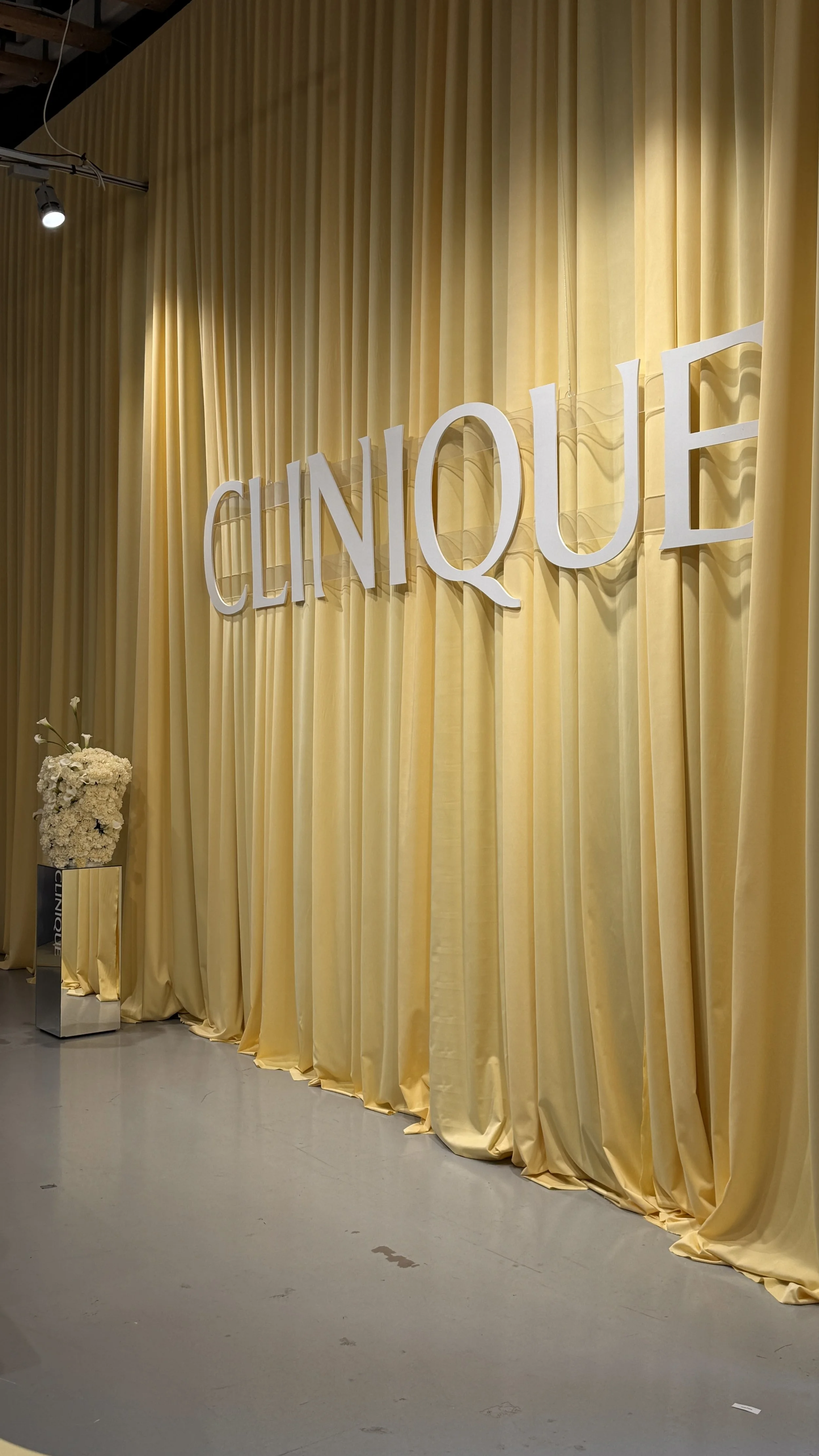 A yellow curtain backdrop with white letters spelling 'CLINIQUE' and a small table with a flower arrangement on the left.