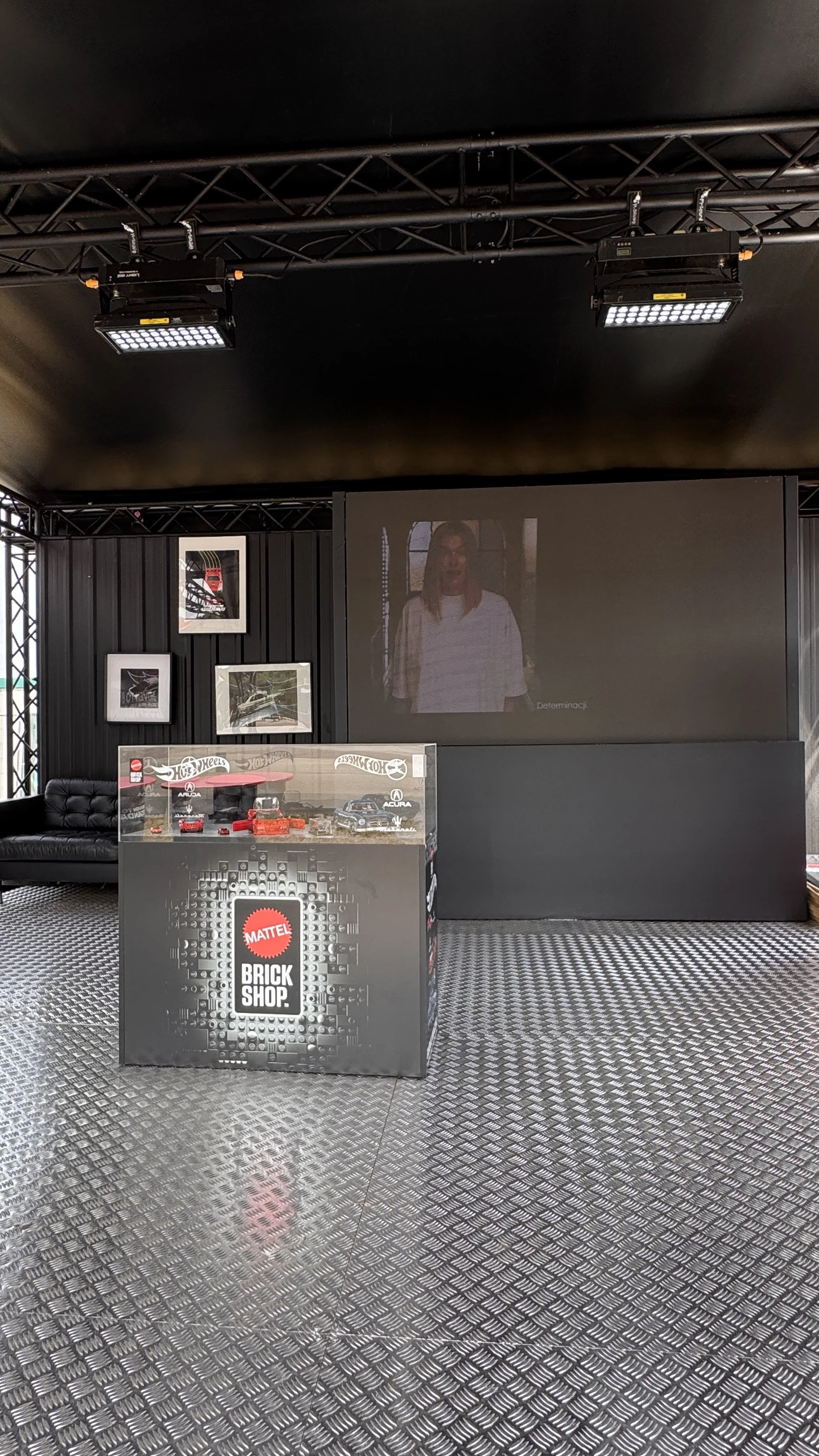 Indoor display area with a large TV screen showing a woman, framed car pictures on the wall, and a counter with Hot Wheels cars in a glass case labeled 'Mattel Brick Shop.'