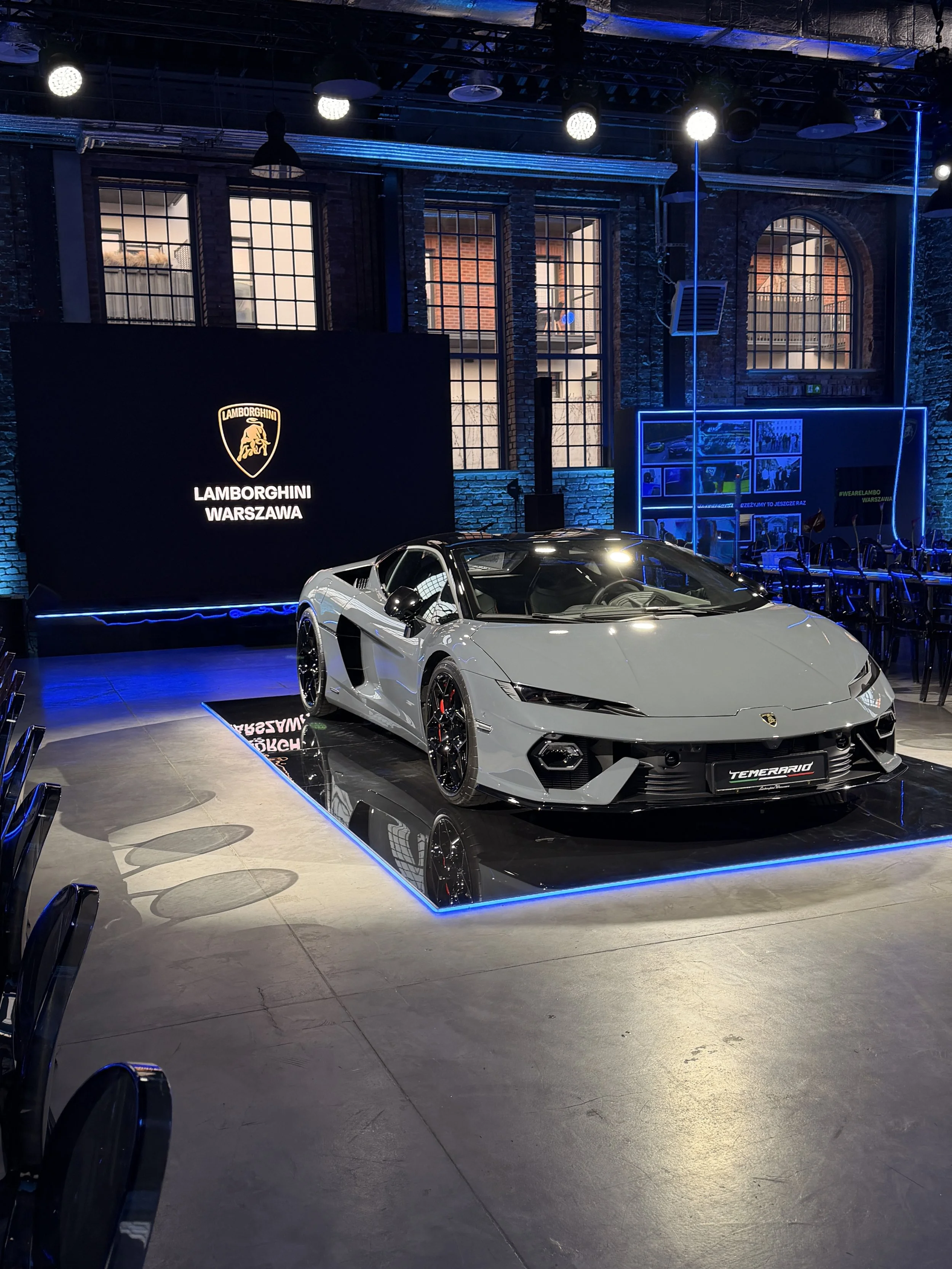 A white Lamborghini sports car on display at an indoor event, with a large Lamborghini logo and the text 'Lamborghini Warszawa' on a black board behind it. The room has brick walls, large windows, and is illuminated with blue and white lights.