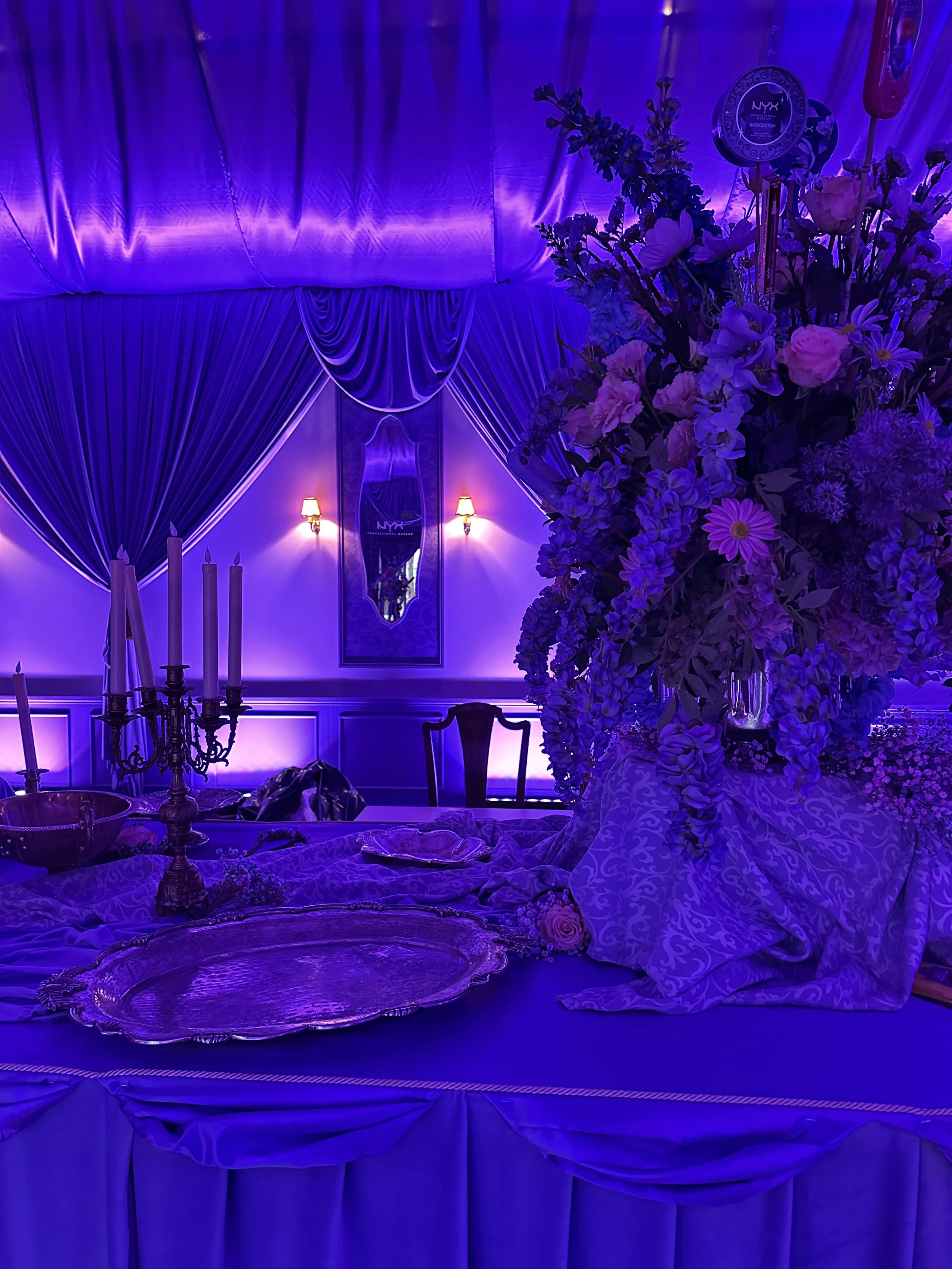 A dimly lit room illuminated with blue and purple lights, decorated with draped velvet curtains on the walls, a large floral arrangement in a vase, an ornate silver tray on the table, and a candelabrum with white candles, suggesting a luxurious or ro