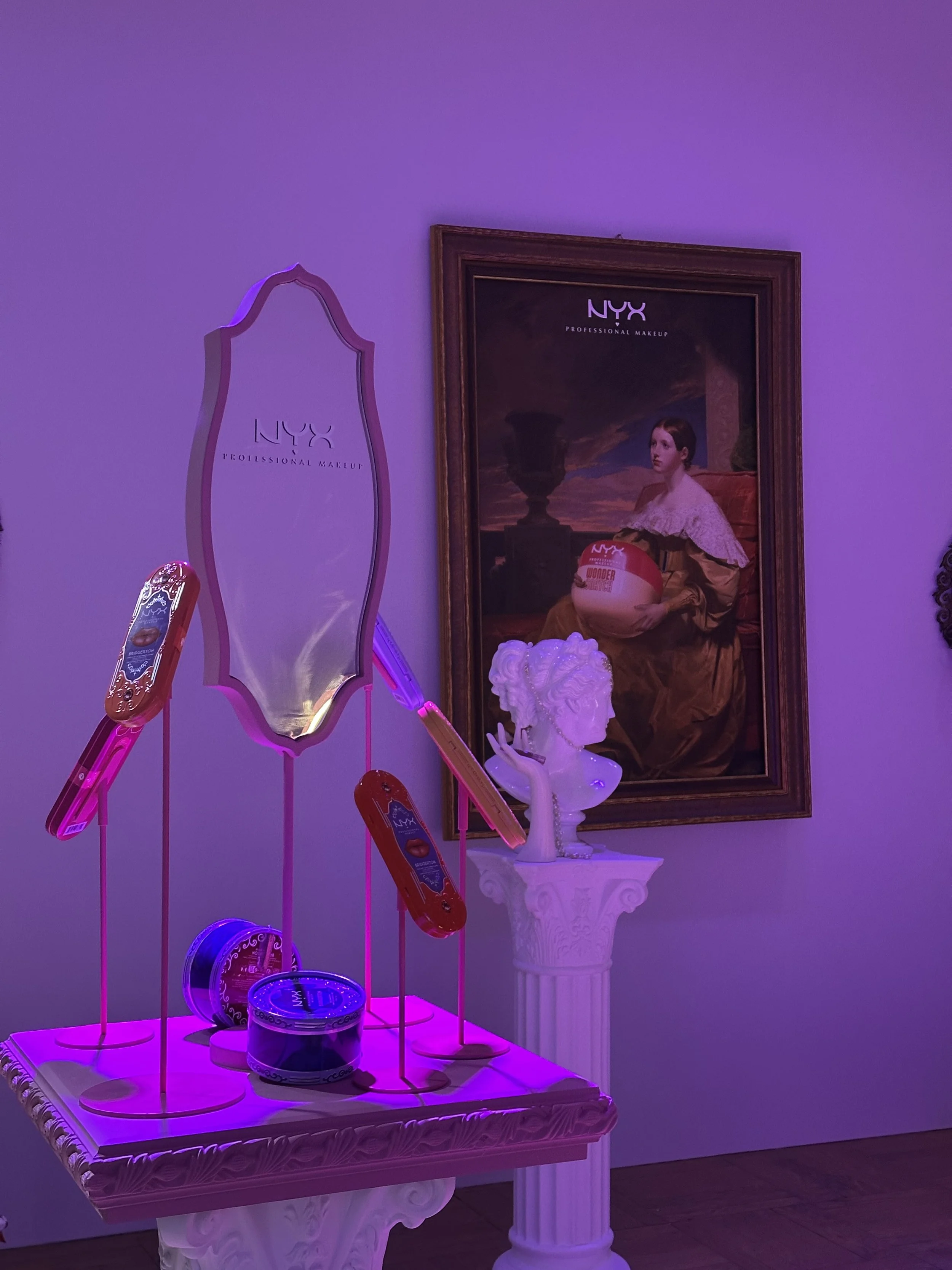 A display of NYX professional makeup products on a decorative table, with a statue bust and a framed painting of a young woman holding a makeup brush in the background, illuminated by purple lighting.