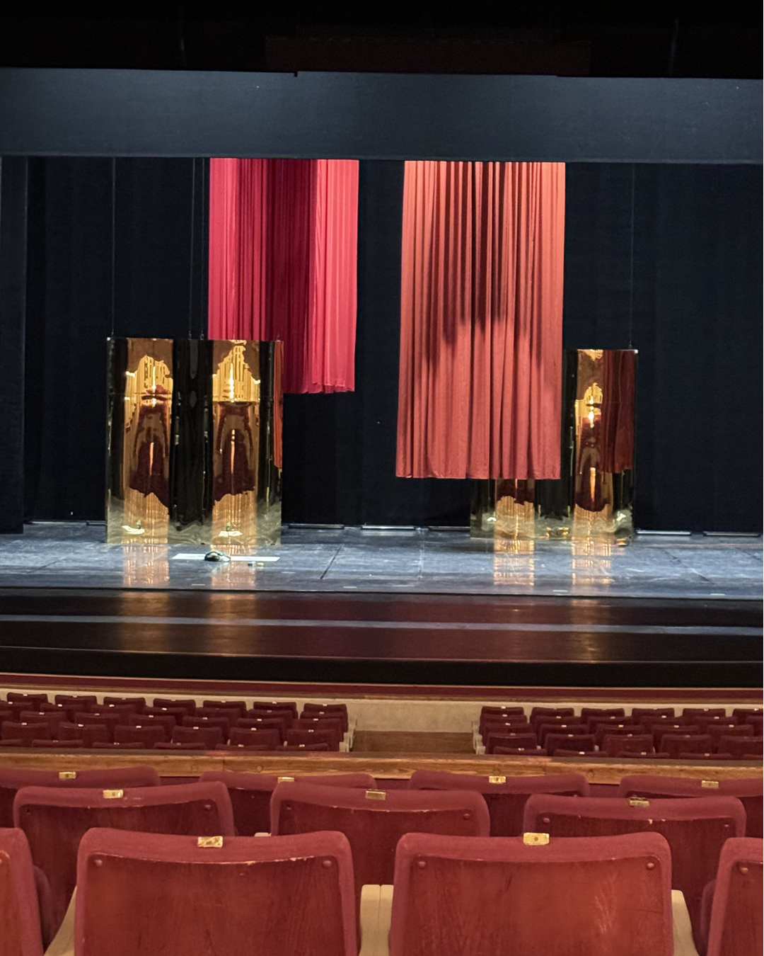 An empty theater stage with red velvet curtains, gold decorative panels reflecting the stage, and rows of red theater seats.