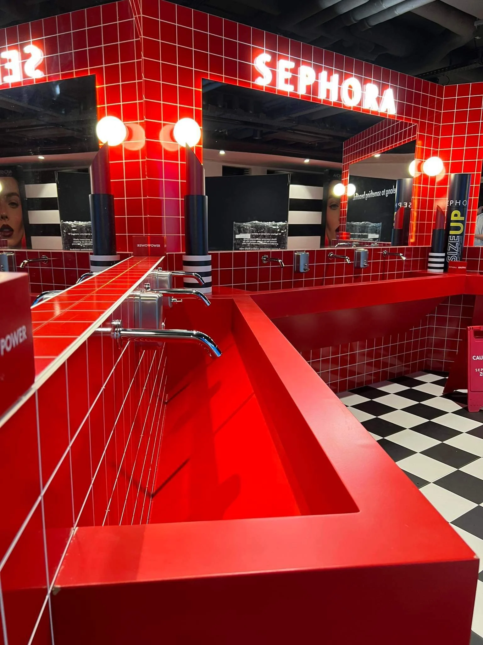 A red tiled counter with two lipstick display stands and a large mirror reflecting a woman’s face in a Sephora store.