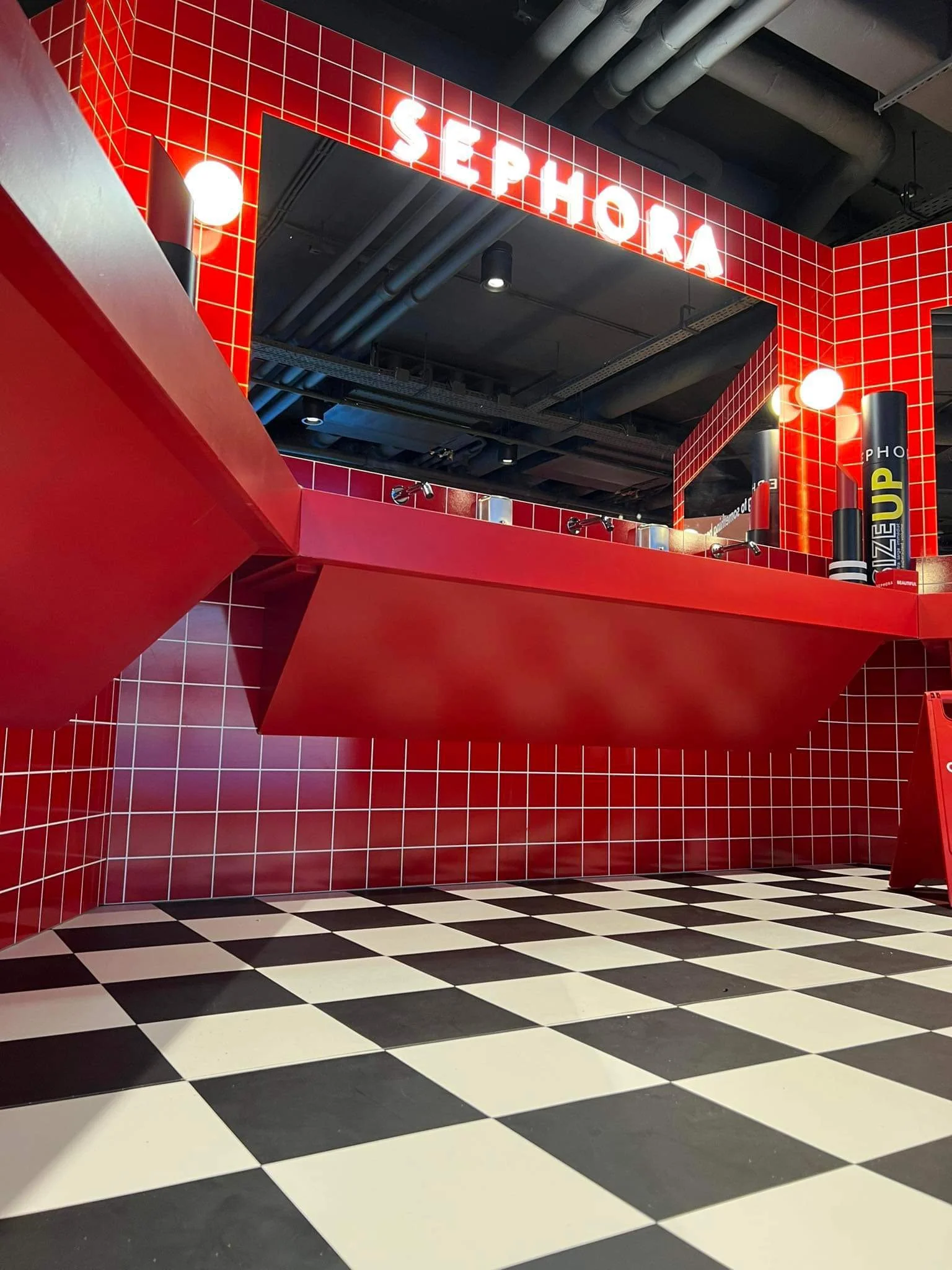 Interior of a store with red wall tiles, a sign that says 'SEPHORA' in bright white letters, and black and white checkered floor. There are red geometric structures and black cylindrical objects on the wall.