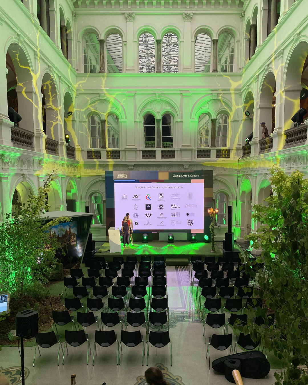 An indoor event in a historic building with high arched windows and ornate architecture. A large screen displays logos of sponsors, and there are chairs arranged for an audience. Green and yellow lighting illuminates the space.
