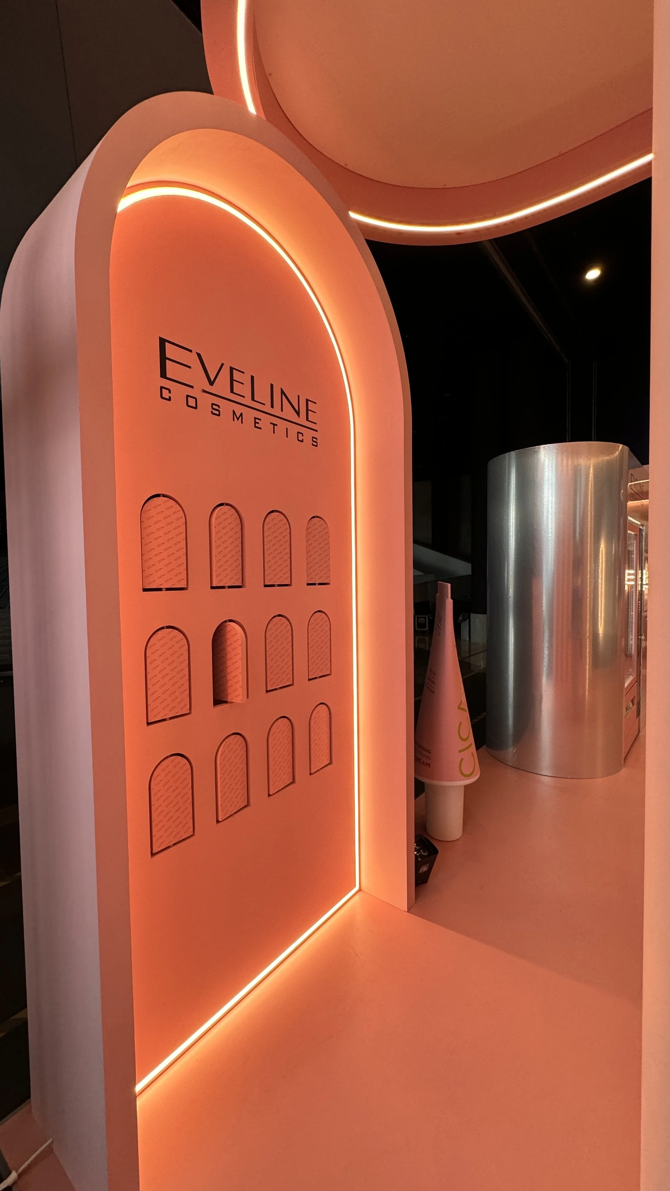 Display stand with the words "Eveline Cosmetics" lit by orange lighting, alongside empty slots for product samples or containers, situated in a retail setting.