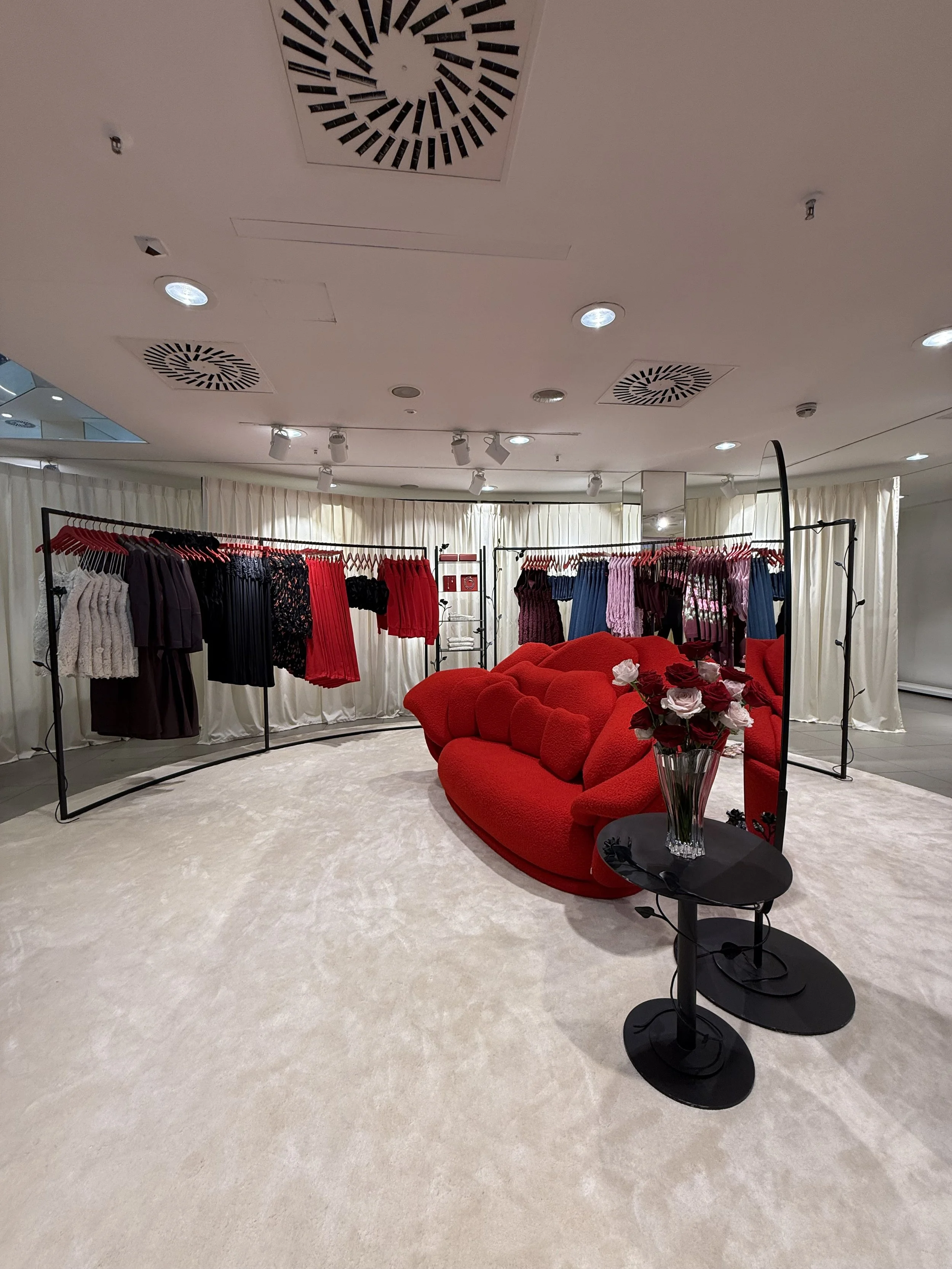 A fashion retail store interior with clothing racks, a red sofa, a black side table with a vase of roses, and ceiling lights.