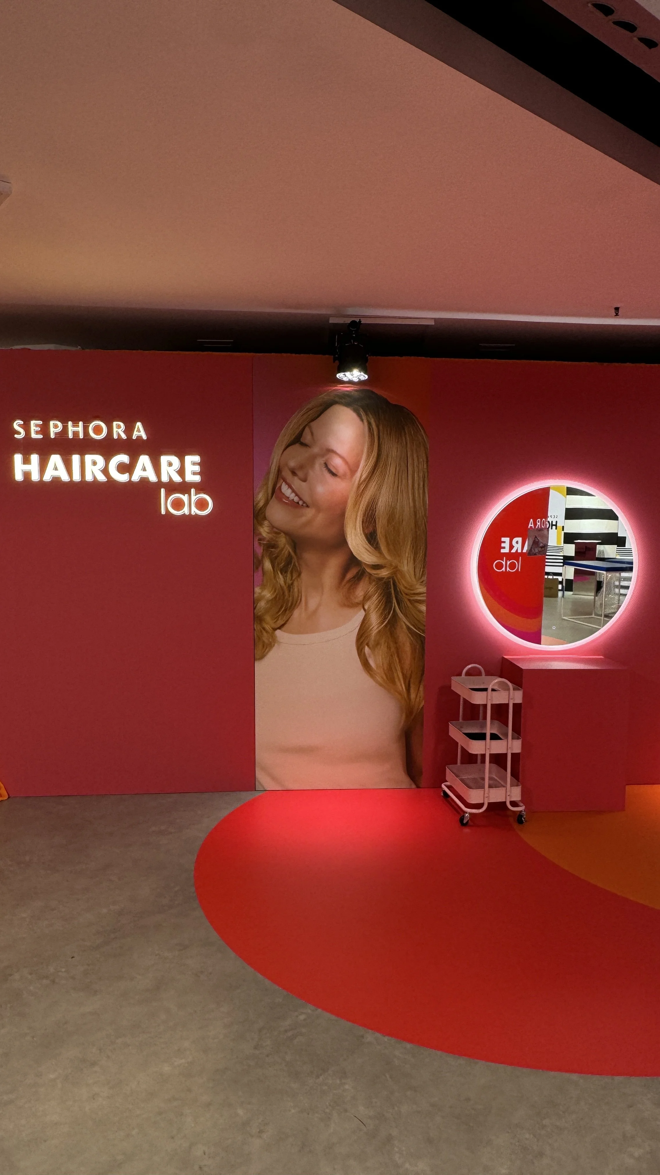 Interior of Sephora Haircare Lab with a large poster of a smiling woman with long red hair, a round illuminated mirror, and a small rolling cart.
