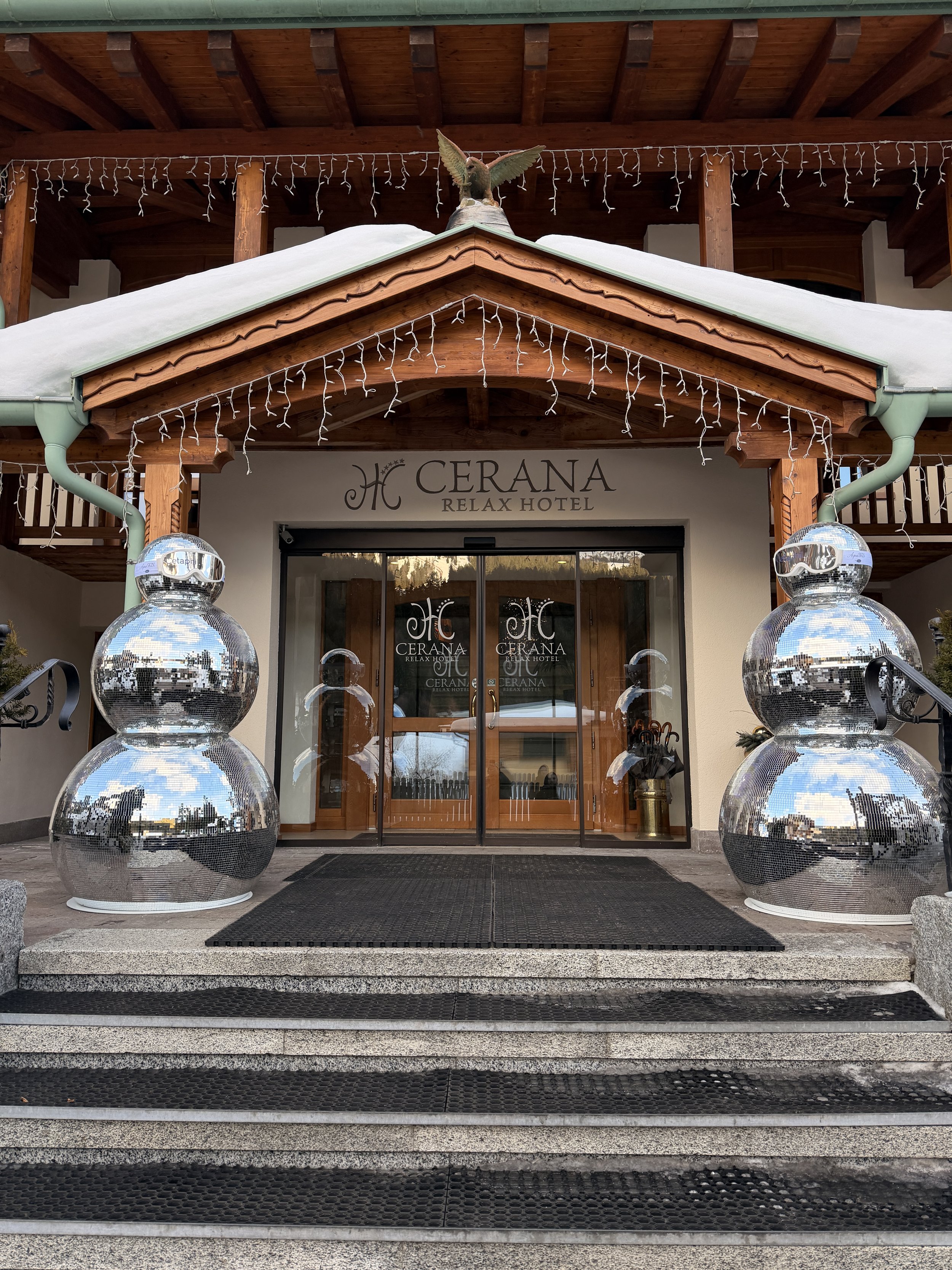 The entrance of Cerana Relax Hotel features a wooden canopy with string lights, a pair of shiny disco ball sculptures on either side of the glass door, and a bird sculpture on the roof corner.