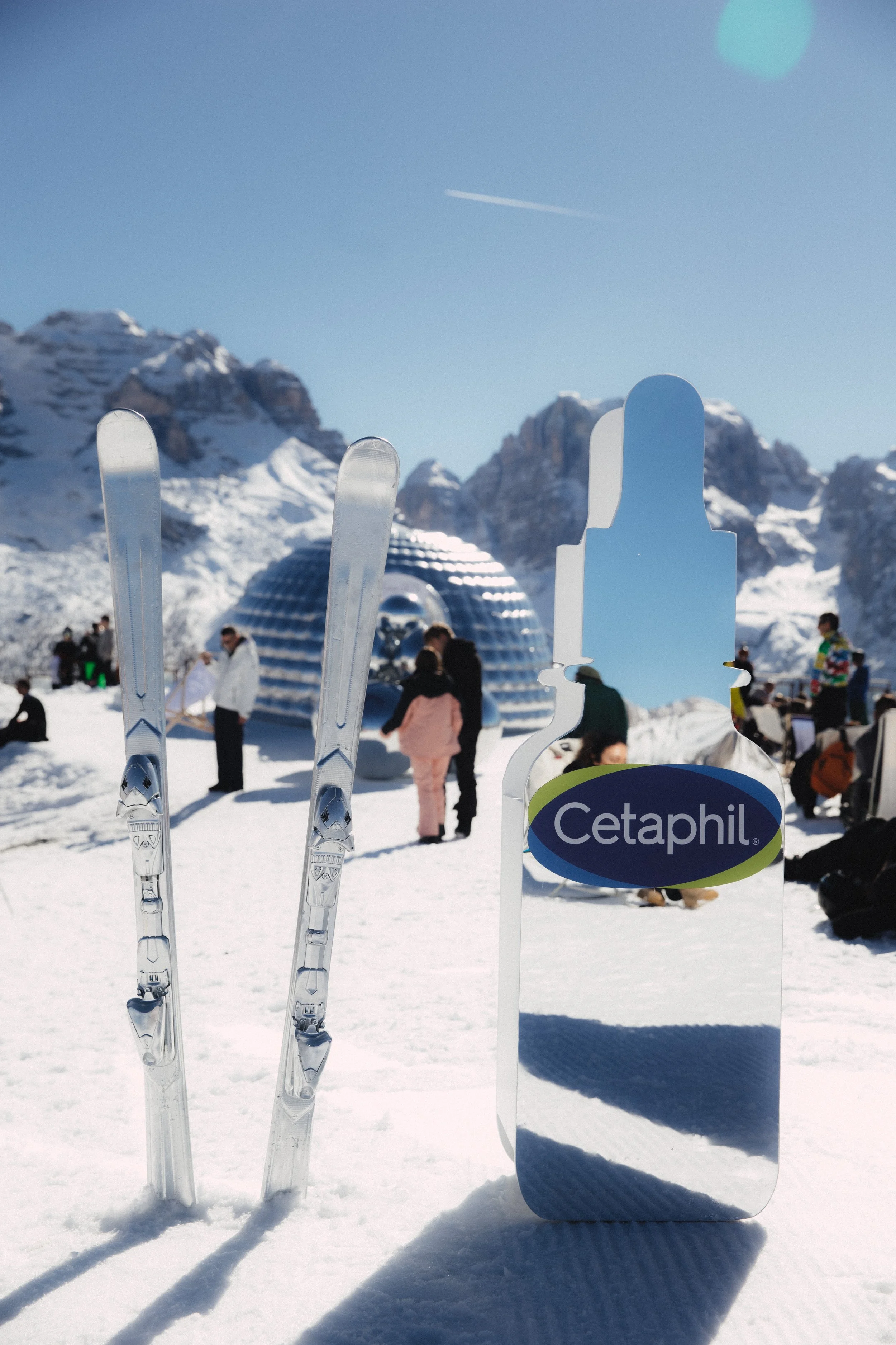 A snowy mountain landscape with skis and ski poles in the foreground. A sign with the Cetaphil logo and people in winter clothing are visible in the background.