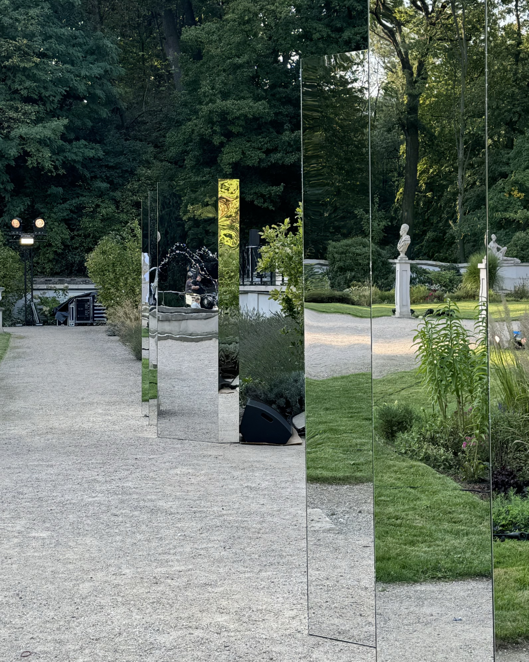 A series of large, reflective mirrors arranged along a gravel pathway in a garden, reflecting trees, statues, and greenery.