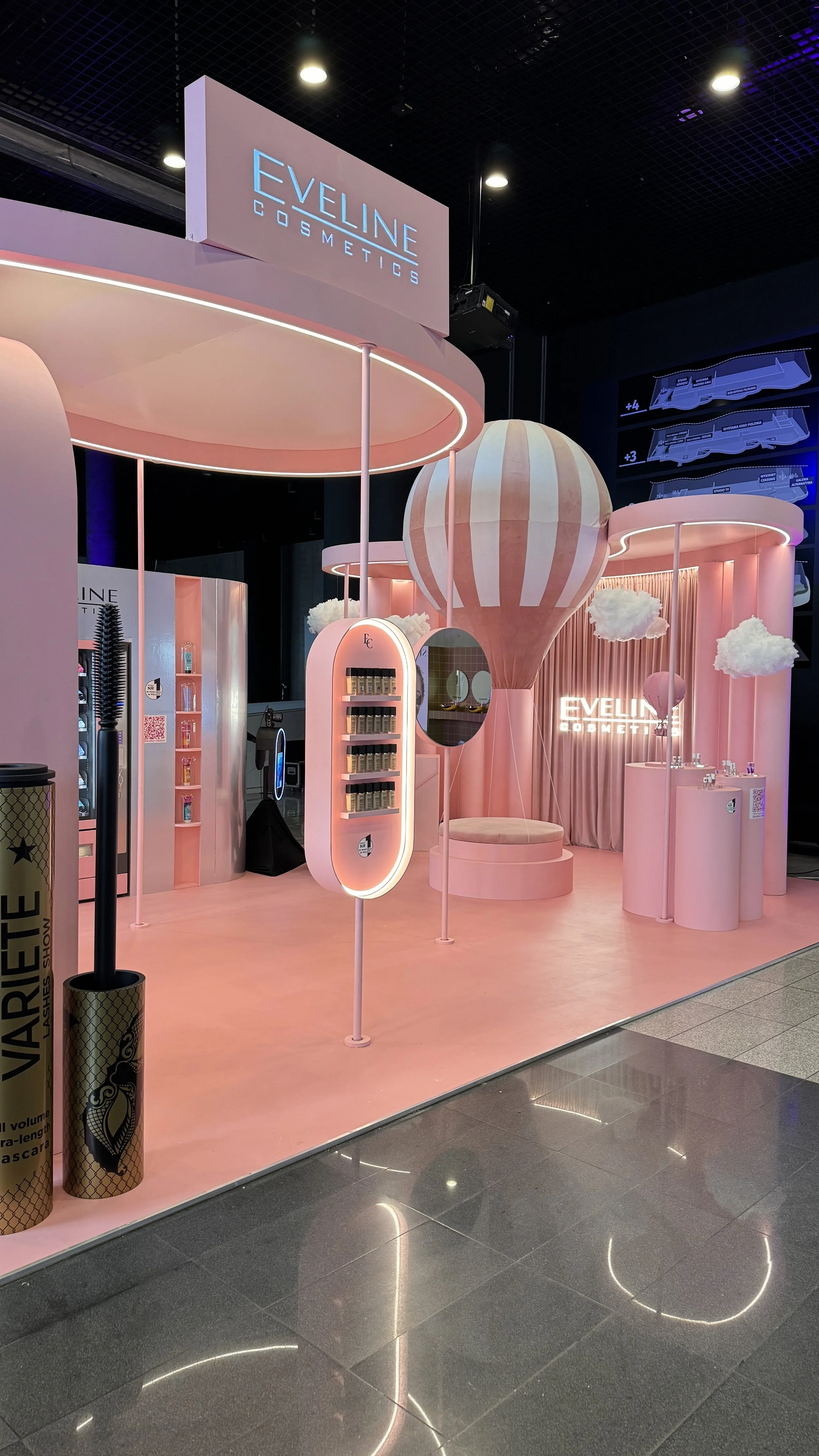 Pink cosmetic booth with cloud, hot air balloon, and circular canopy decorations, displaying Eveline Cosmetics branding at an indoor event, with floor and ceiling reflections visible.