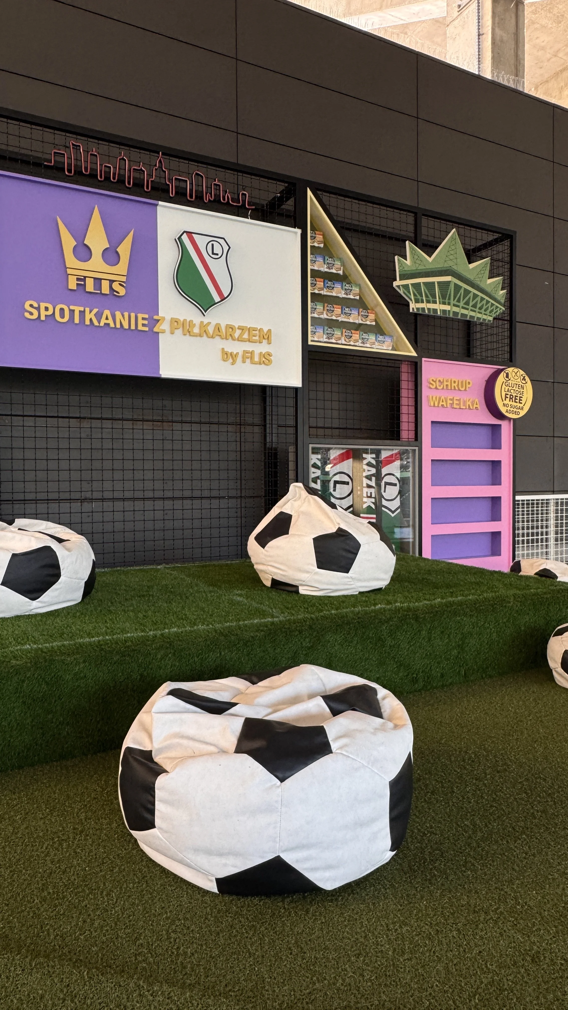 Indoor area with soccer-themed decor, large bean bags resembling soccer balls on artificial grass, and colorful signs, including one that reads 'Spotkanie z Piłkarzem'.