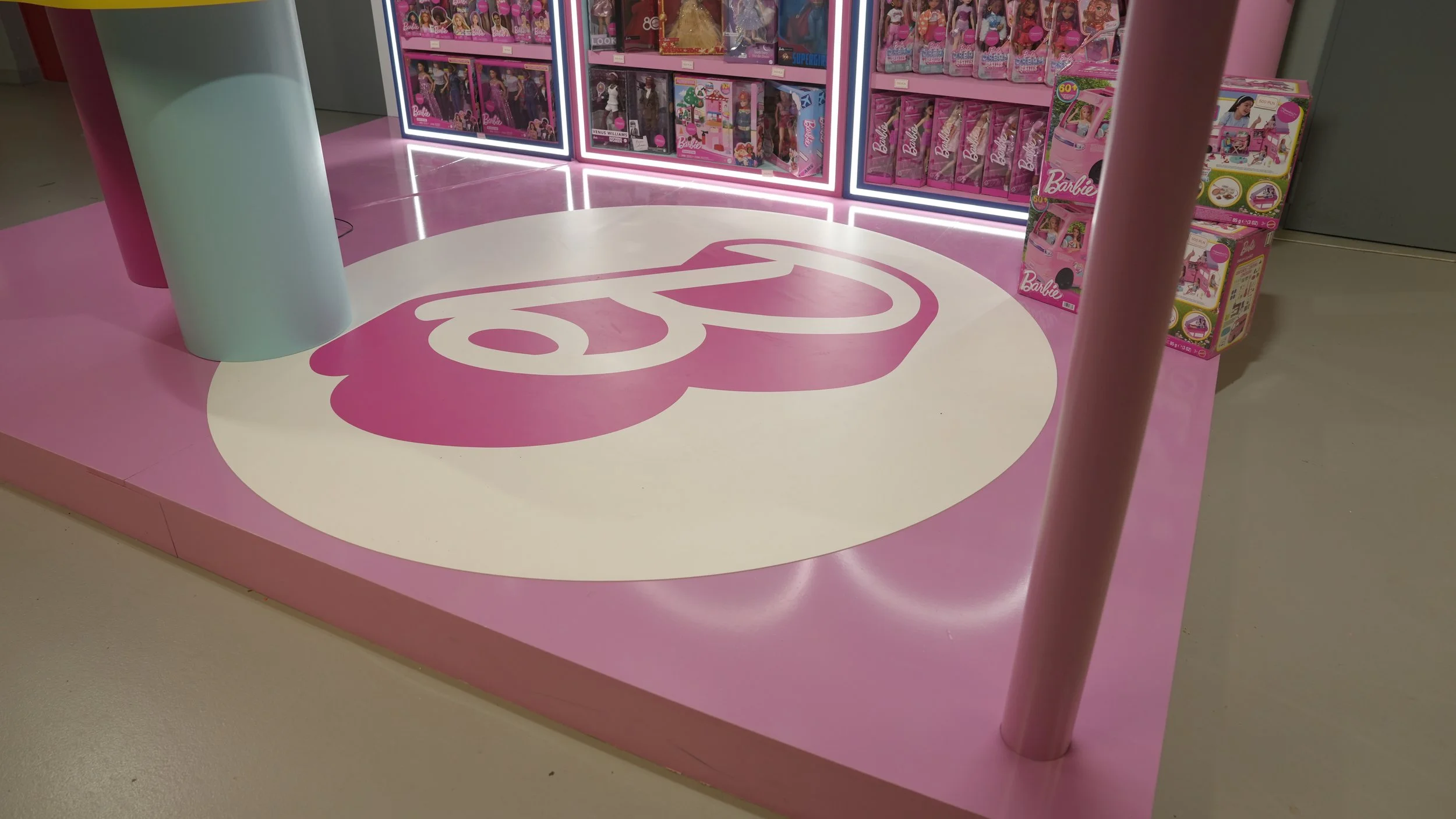 Display of Barbie dolls on pink shelves at a toy store, with a large pink and white circular logo on the floor.