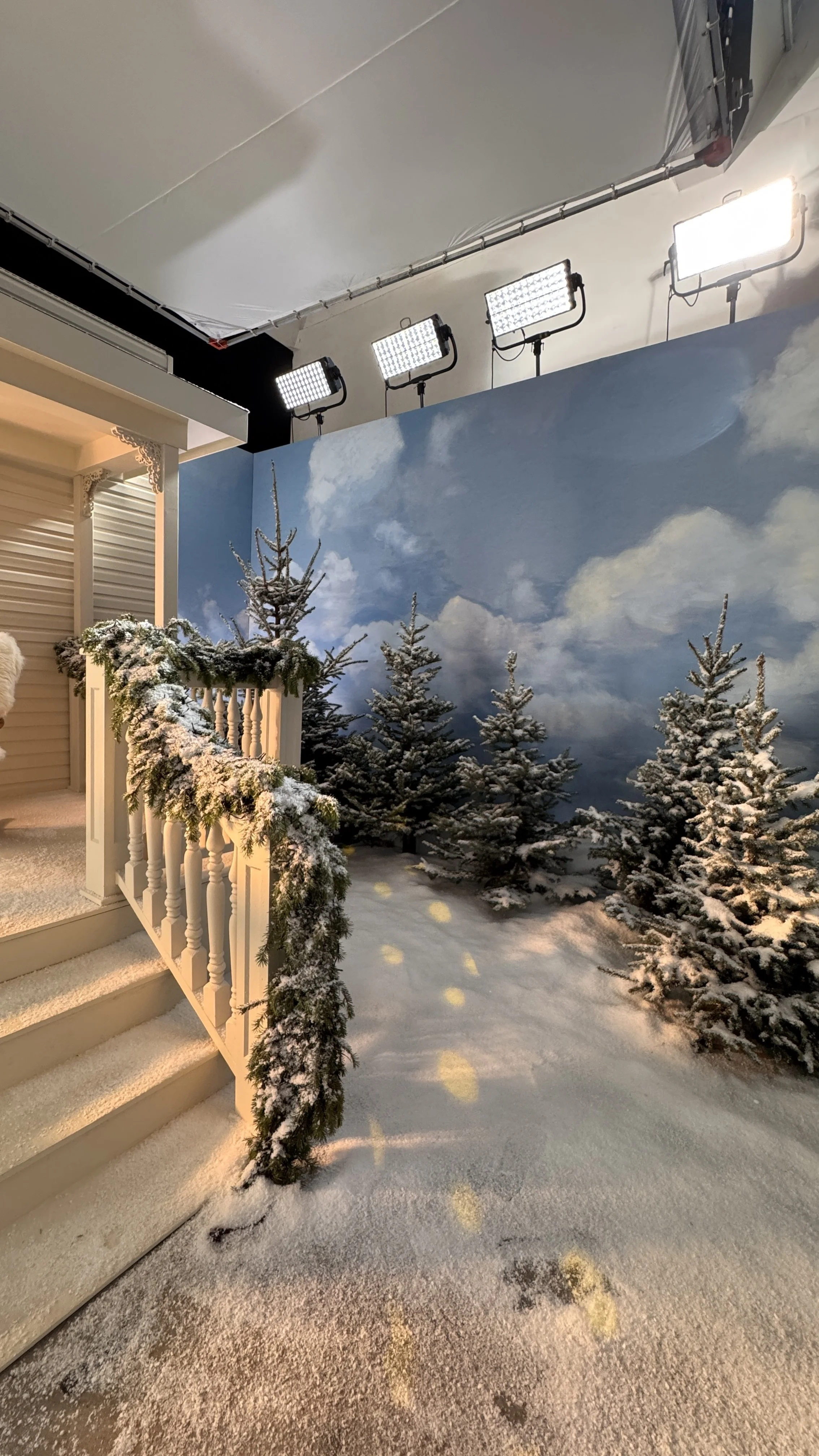 Indoor scene with snow-covered Christmas trees and decorated stairs with Christmas garland, against a painted sky mural with clouds. Overhead, bright studio lights illuminate the scene.