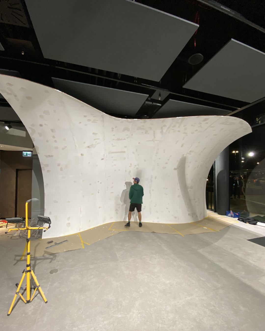 A person practicing rock climbing on an indoor climbing wall with a distinctive curved design. The wall is white and textured, with some markings and scuff marks. The person is wearing a green hoodie, black shorts, and a cap. There are some climbing 