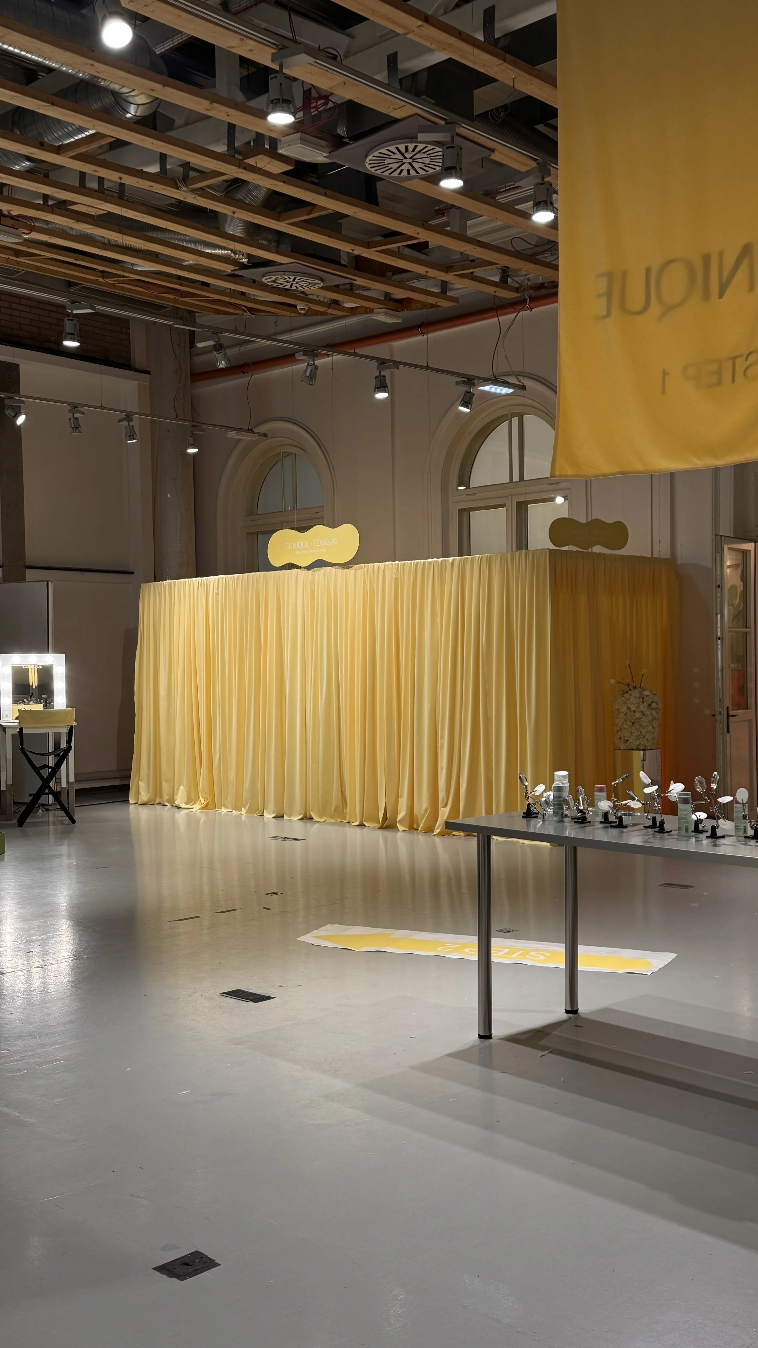 An indoor space with a yellow curtain, a table with jewelry display stands, and a vanity mirror with lights. The ceiling shows exposed wooden beams and ductwork.