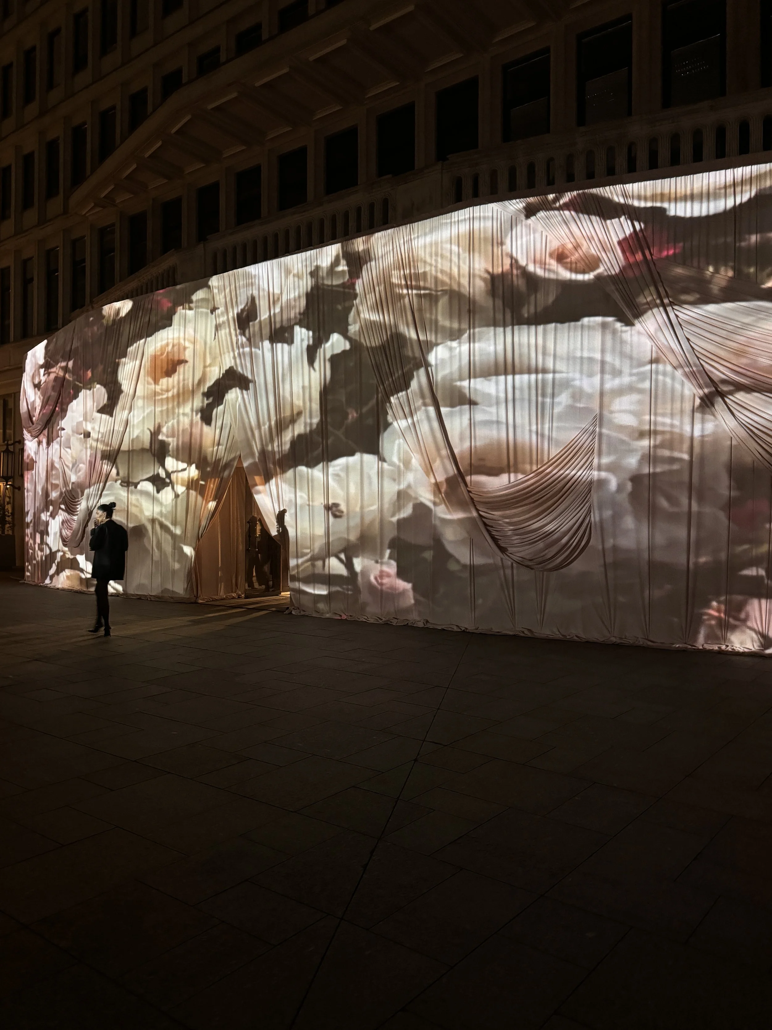 Nighttime outdoor scene with a building in the background and a large, illuminated fabric display with floral patterns and draped fabric, along with a person standing in front.