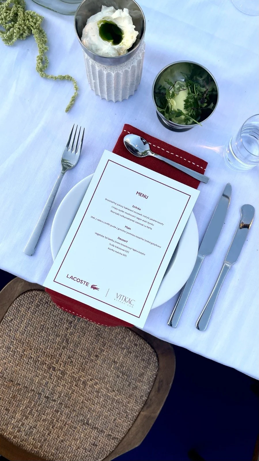 A table set for a meal with a white plate, silver fork, knife, and spoon, a red napkin, and a menu. There are two glasses, one filled with water and the other with greenery and herbs. A glass with whipped cream and a green drizzle on top is also on t