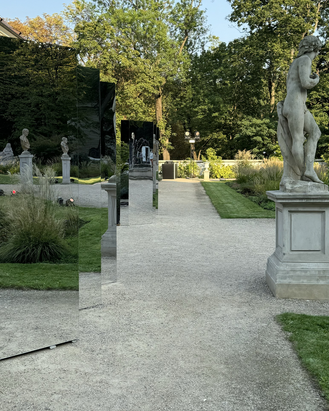 Stone sculptures of classical figures and mirrored installation reflecting trees and statues in a garden setting, surrounded by greenery and gravel pathways.