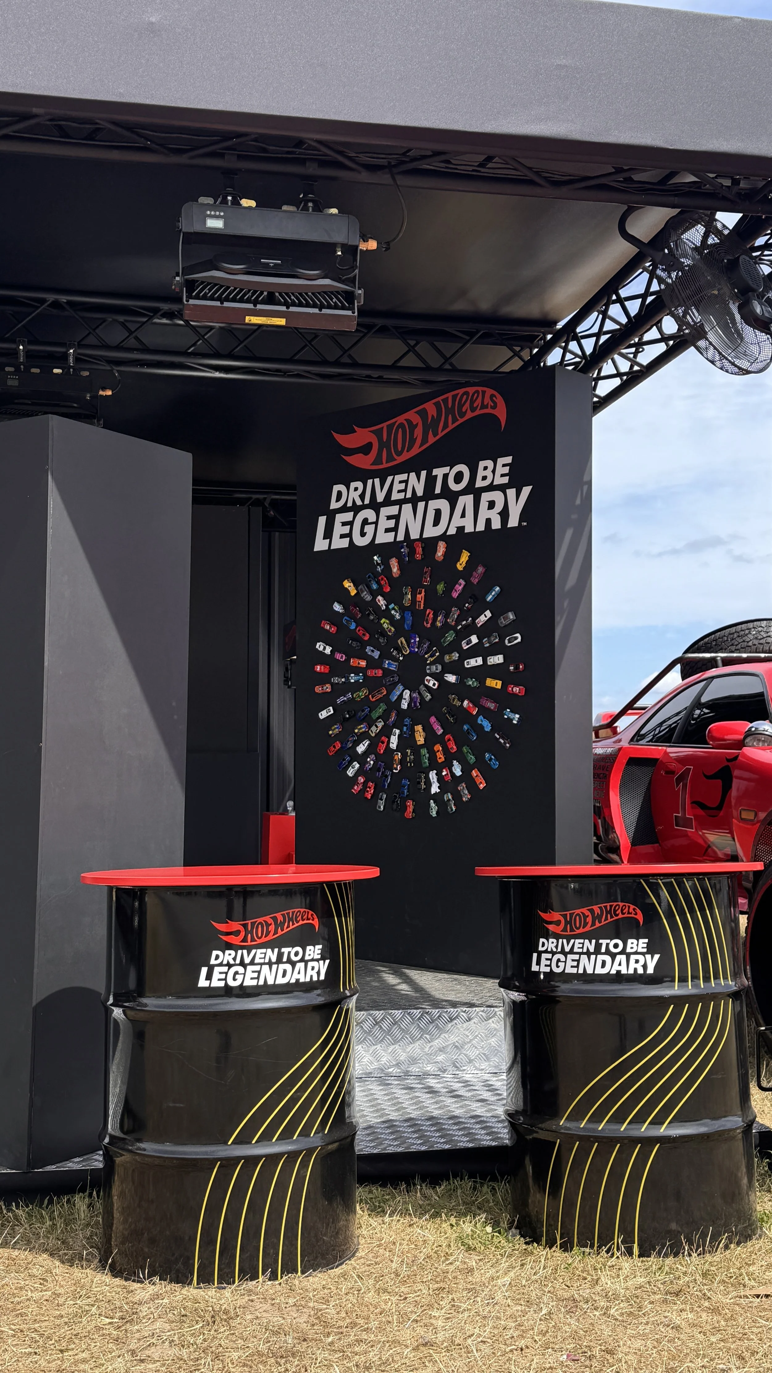 A Hot Wheels display at an outdoor event, featuring a black structure with a collection of small, colorful toy cars arranged in a spiral pattern, and Hot Wheels branding with the slogan 'Driven to be Legendary' on black barrels in the foreground.