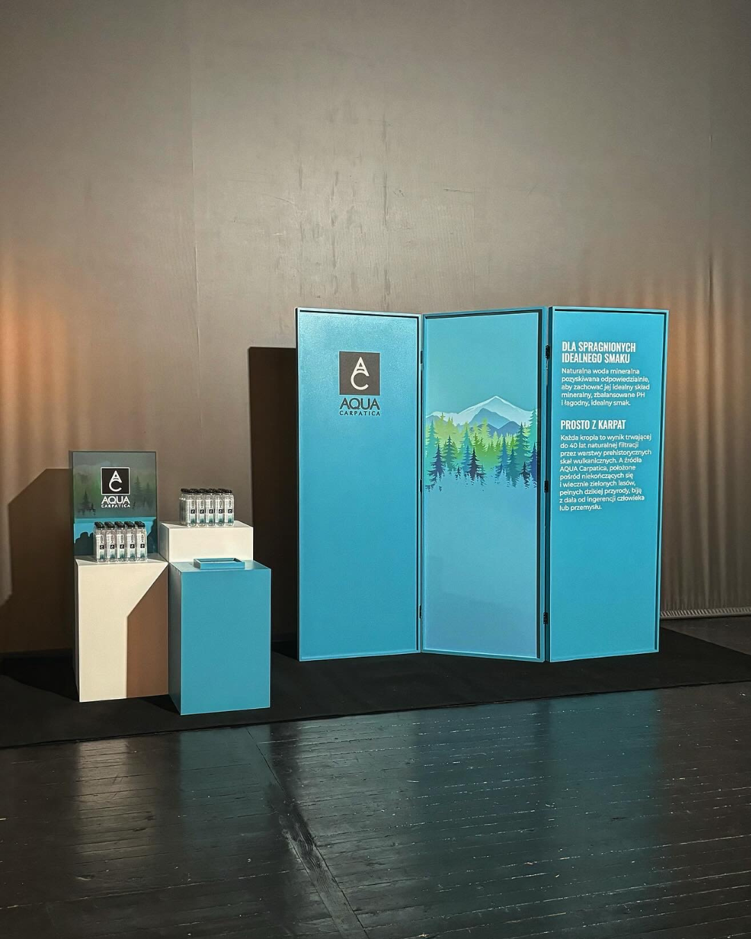 A promotional display for Aqua Carpatica mineral water, featuring a blue folding screen with mountain and forest illustrations, and bottles of water on white and blue stands.