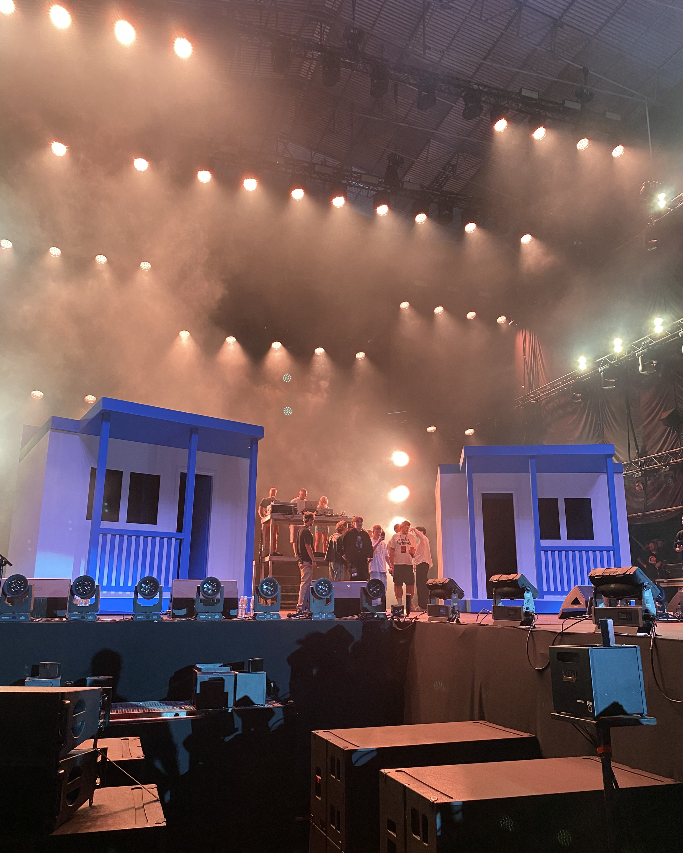Stage setup with colorful lighting, blue structures resembling houses, and a group of people preparing for a performance or event in a large indoor venue.