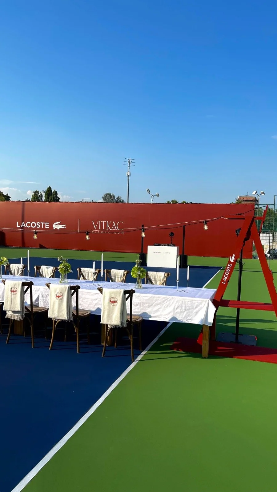 Outdoor tennis court with a table set for a meal, decorated with small flower arrangements, and strings of lights hung above. A red wall with logos for Lacoste and Vitkac in the background.