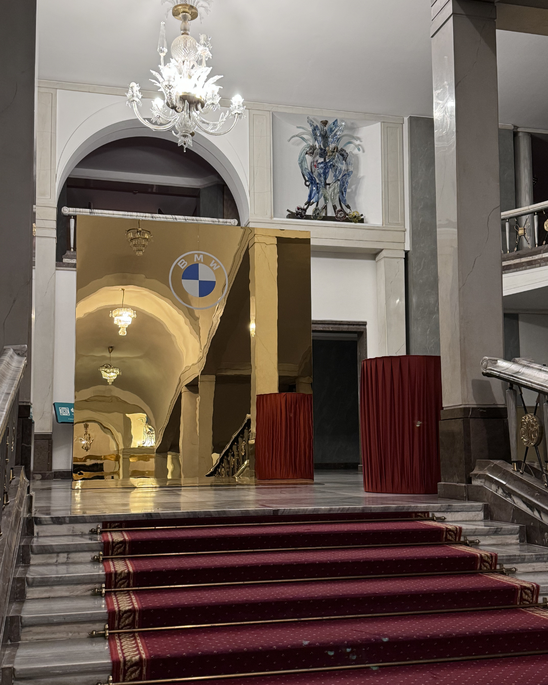 Interior entrance hall with marble stairs, red carpet, chandelier, a large gold mirror, and decorative sculptures