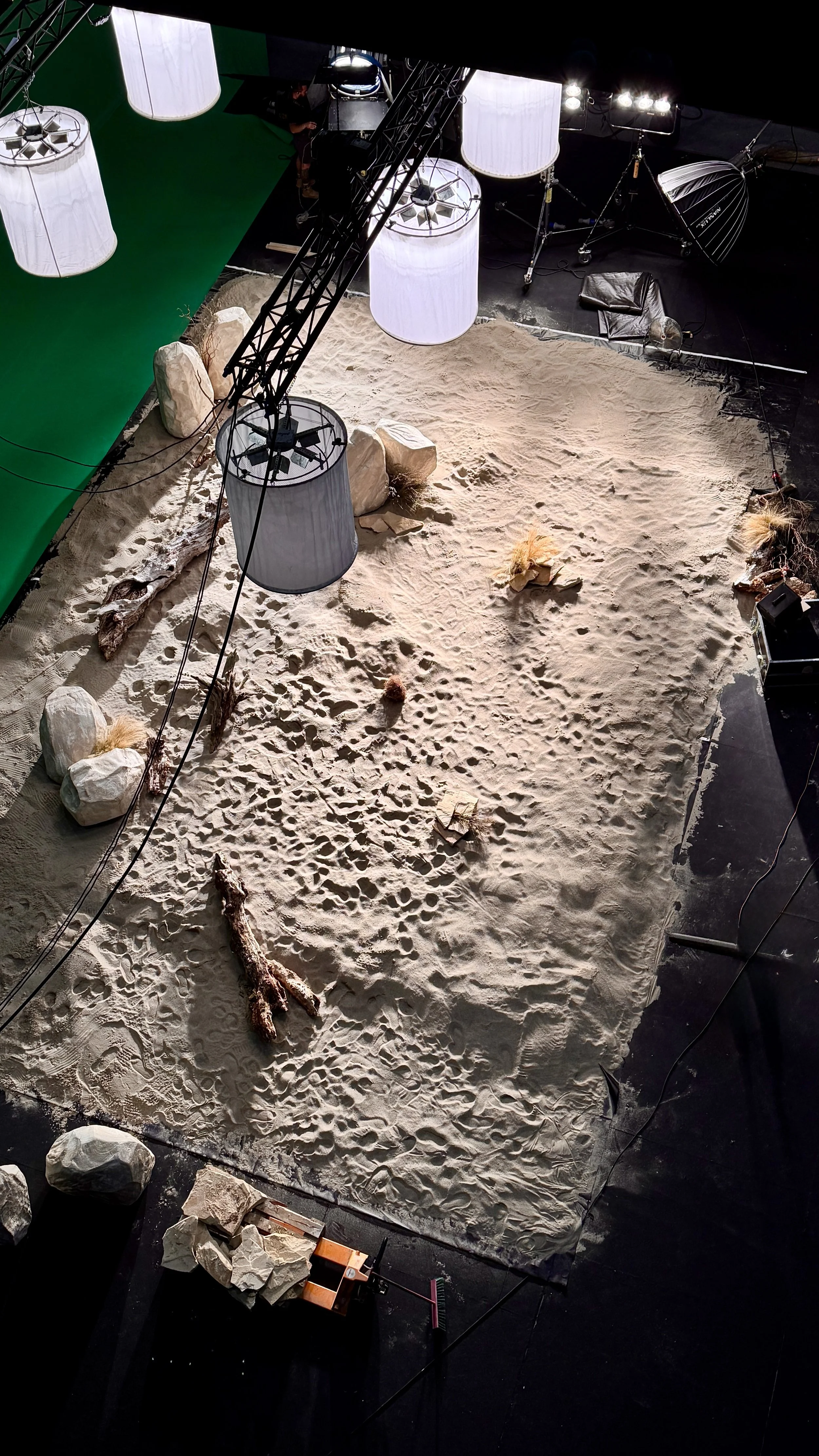 A behind-the-scenes view of a film or television set showing a sandy floor with rocks and driftwood, large hanging white lights, green screen on the left, studio equipment, and a crew member working on the set.