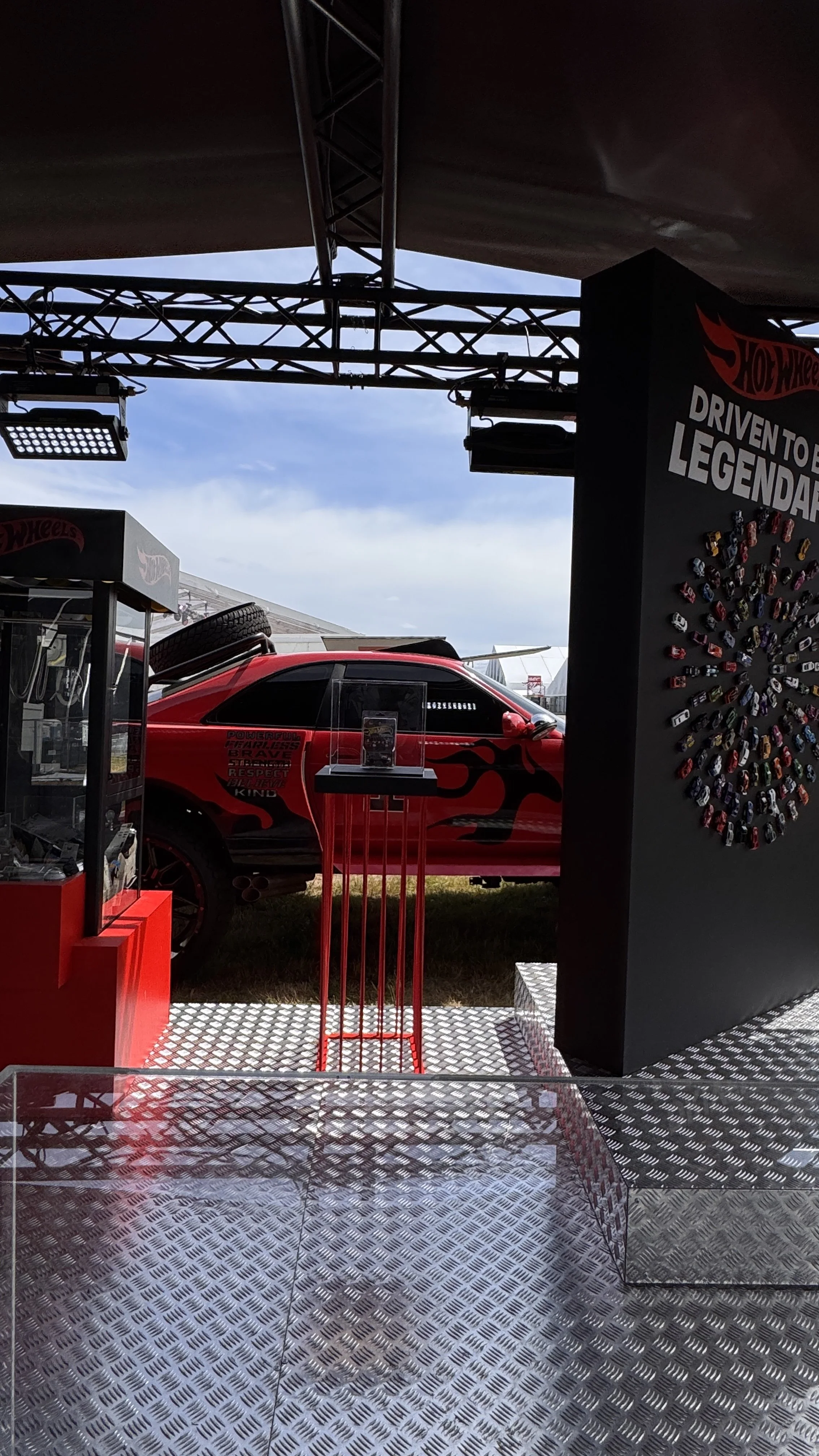 A red race car with flame decals on the side displayed at an outdoor event, with signs and display stands around it and a partly cloudy sky in the background.