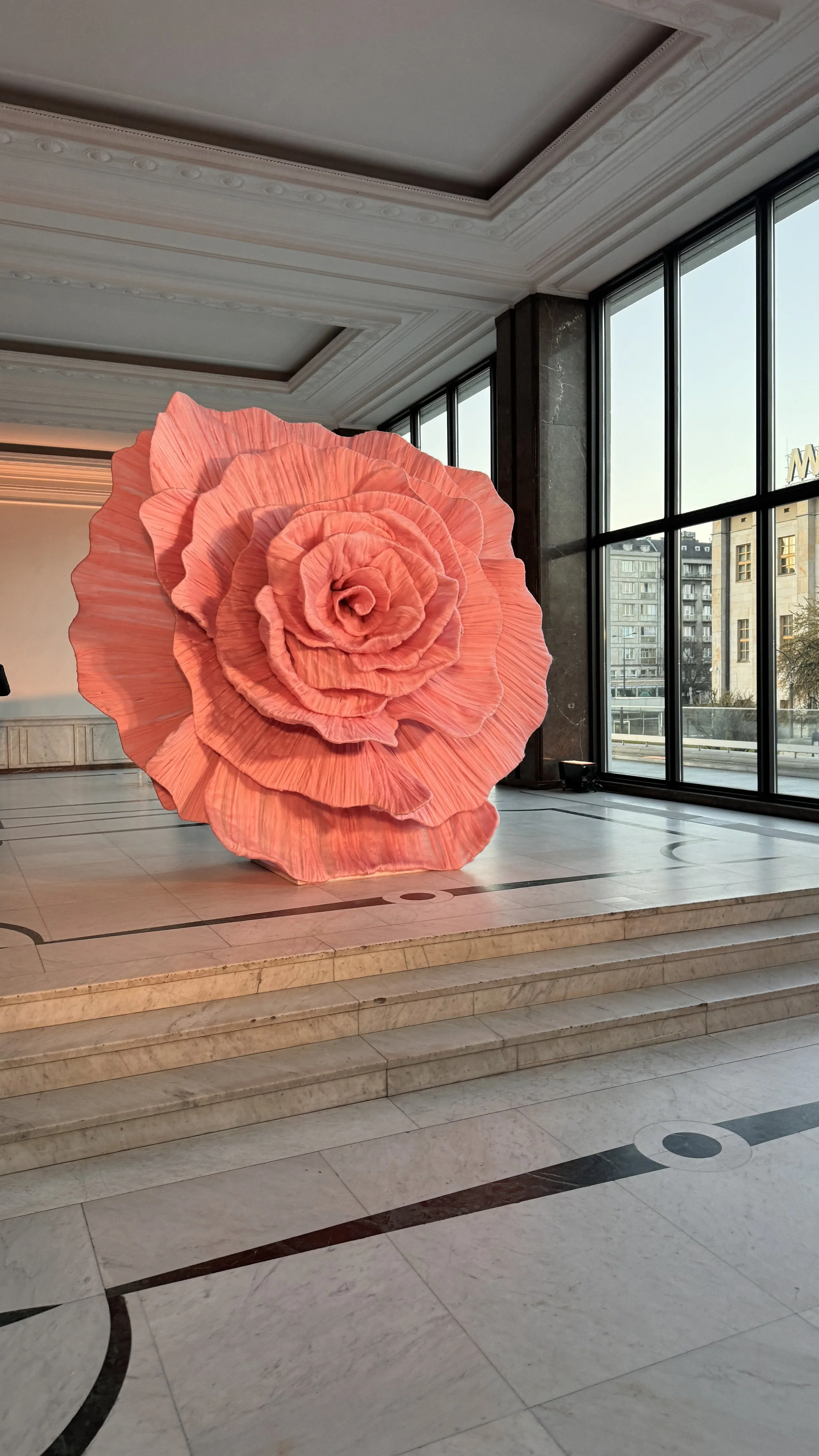 A large, pink, paper flower sculpture resembling a rose inside a spacious room with marble floors, white ornate ceiling molding, and tall windows showing a cityscape outside.