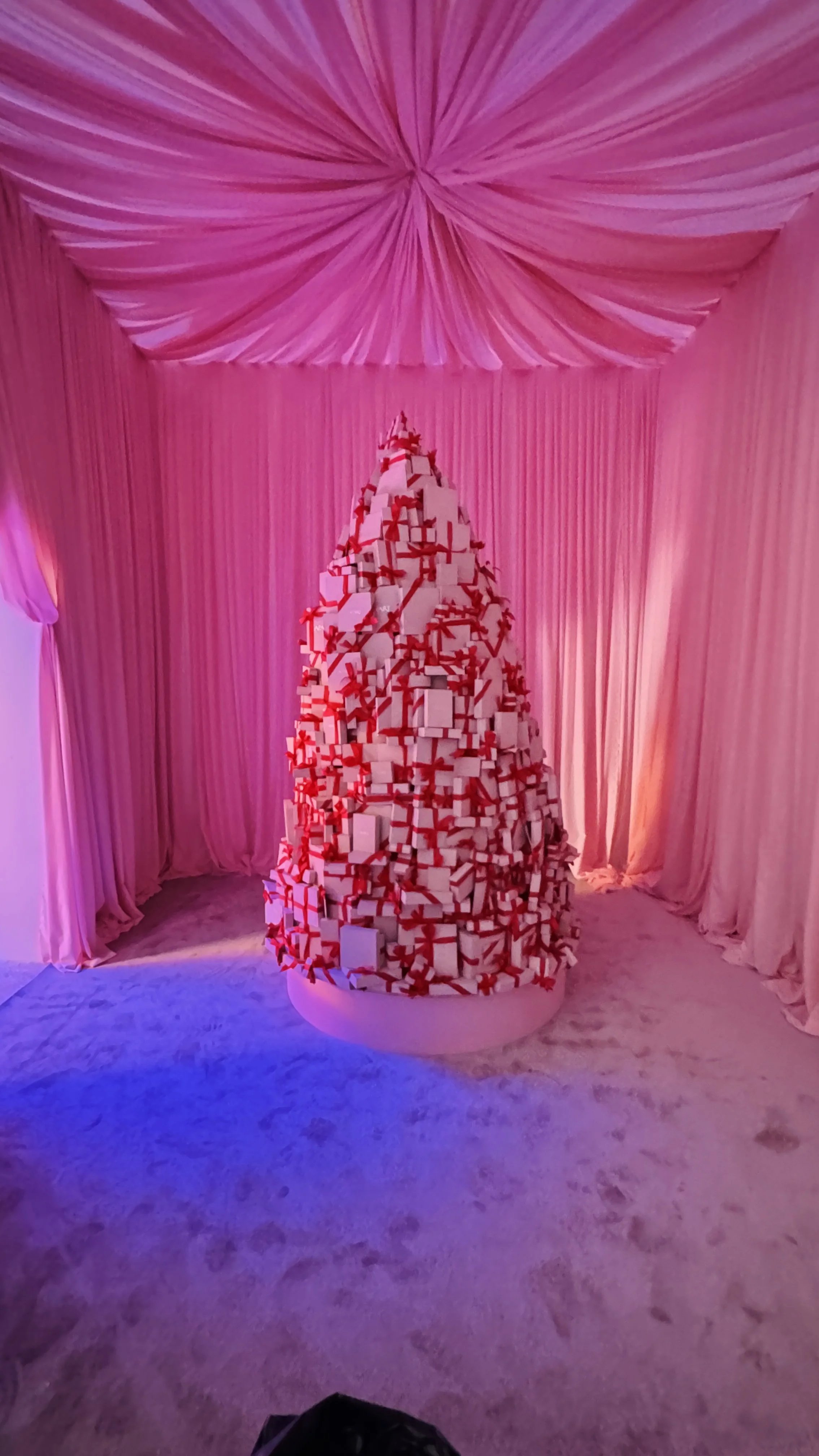 A Christmas tree made of many white gift boxes with red ribbons, set on a circular platform in a room with pink drapery and ceiling decoration, illuminated with pink and purple lighting.