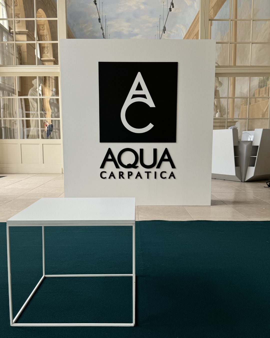 A black and white sign reading 'Aqua Carpatica' with a large 'AC' logo above, positioned on a white display board in a modern indoor space with glass and decorative elements in the background.