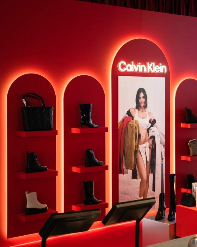 Calvin Klein store display with red background, illuminated arches, black and white boots on red shelves, and a large poster of a woman in casual clothing.