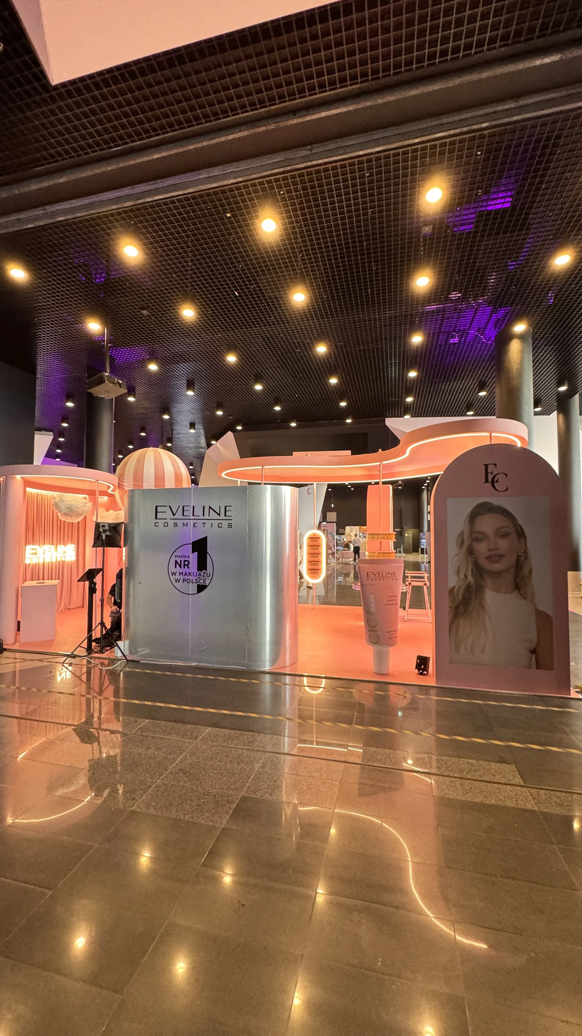 Indoor promotional display for Eveline Cosmetics with pink and orange lighting, featuring a large image of a woman with blonde hair, a giant skincare product, and branded signage.