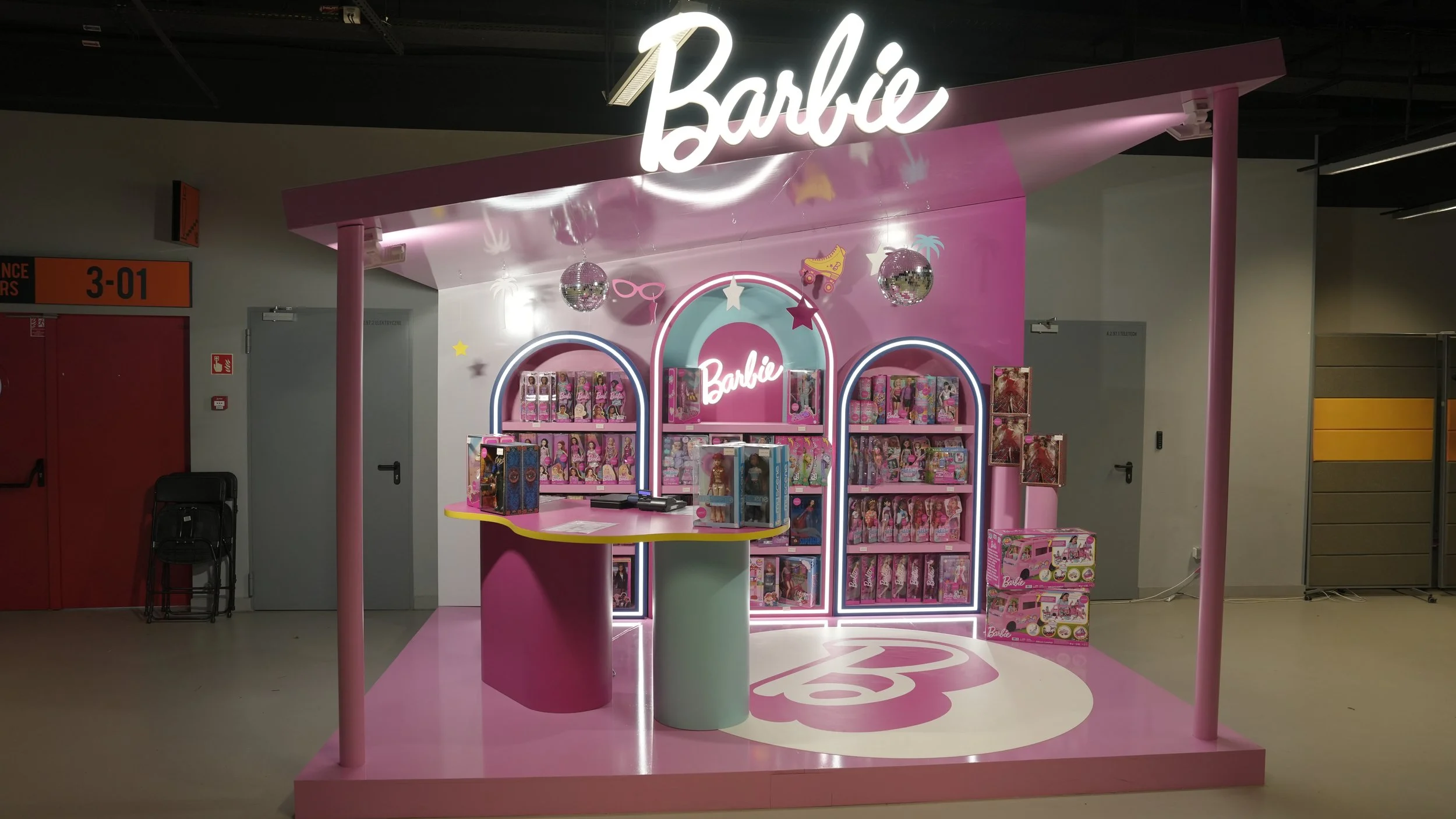 Pink Barbie merchandise kiosk with various dolls and accessories on display, topped with a neon 'Barbie' sign, in an indoor shopping area.