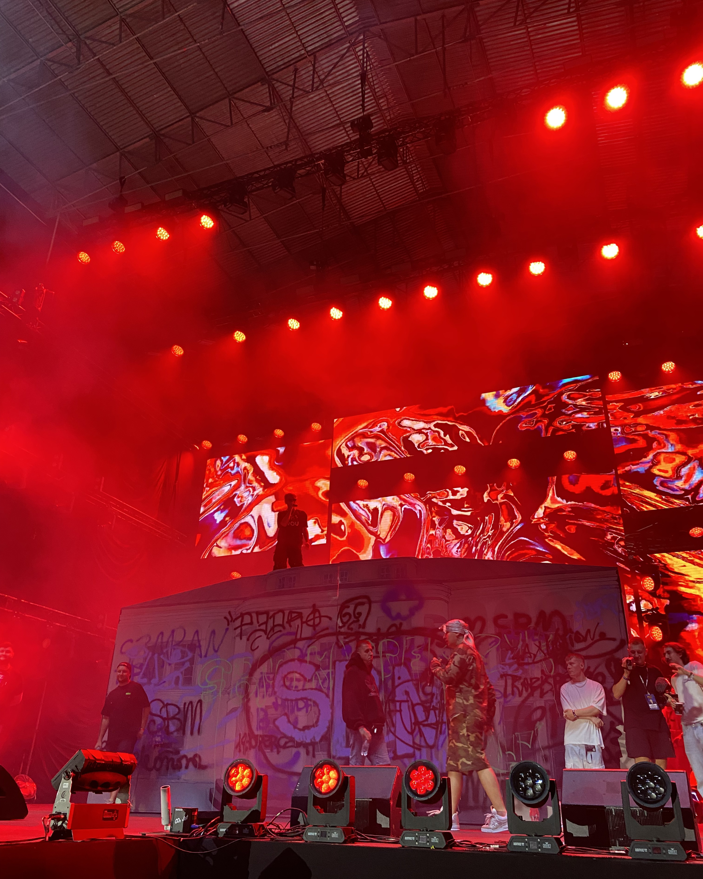 People on stage with a graffiti-covered backdrop, illuminated by red stage lights, at a concert or event.