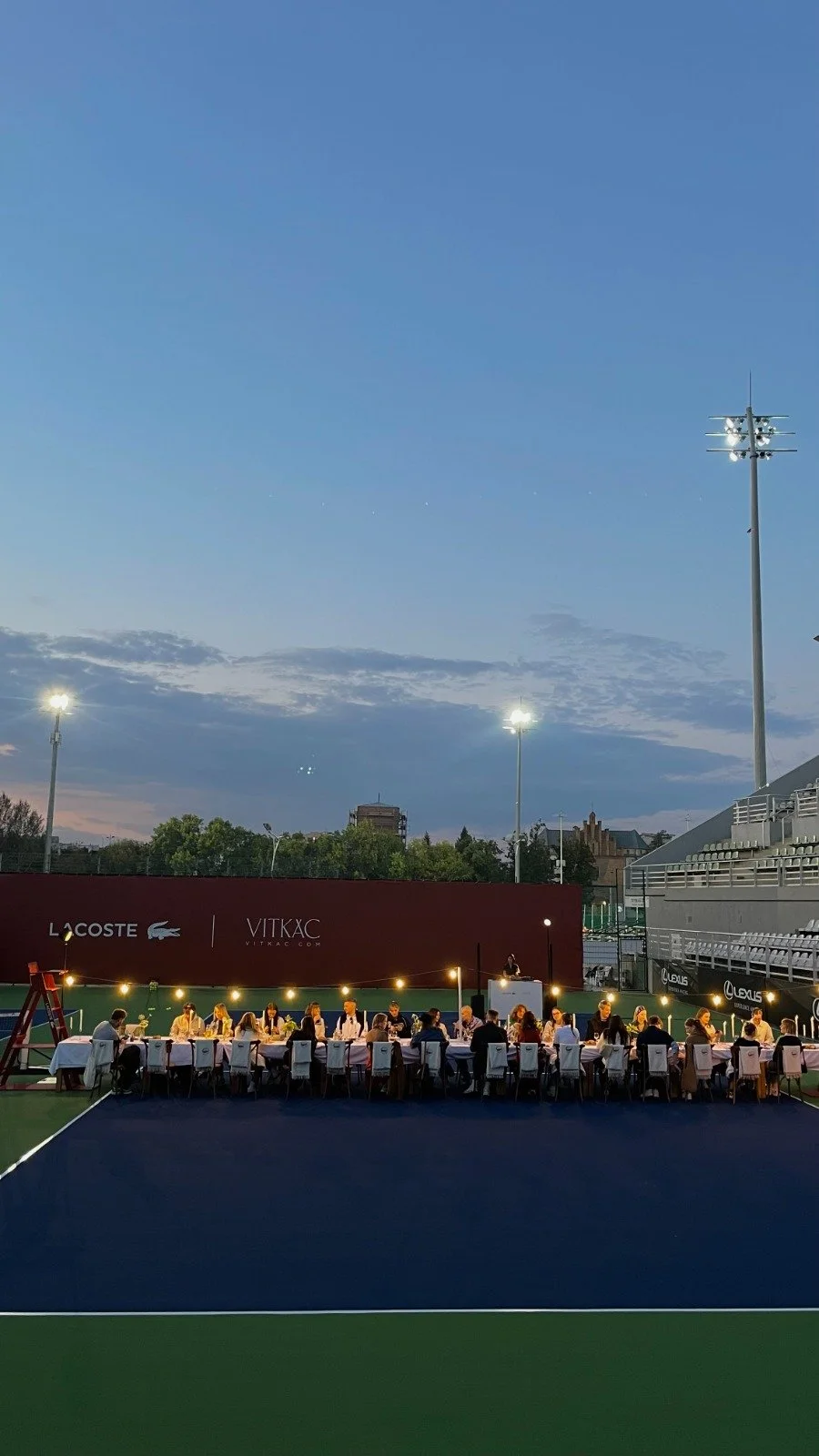 People dining at a long table on a tennis court during twilight, illuminated by string lights, with stadium lights and buildings in the background.