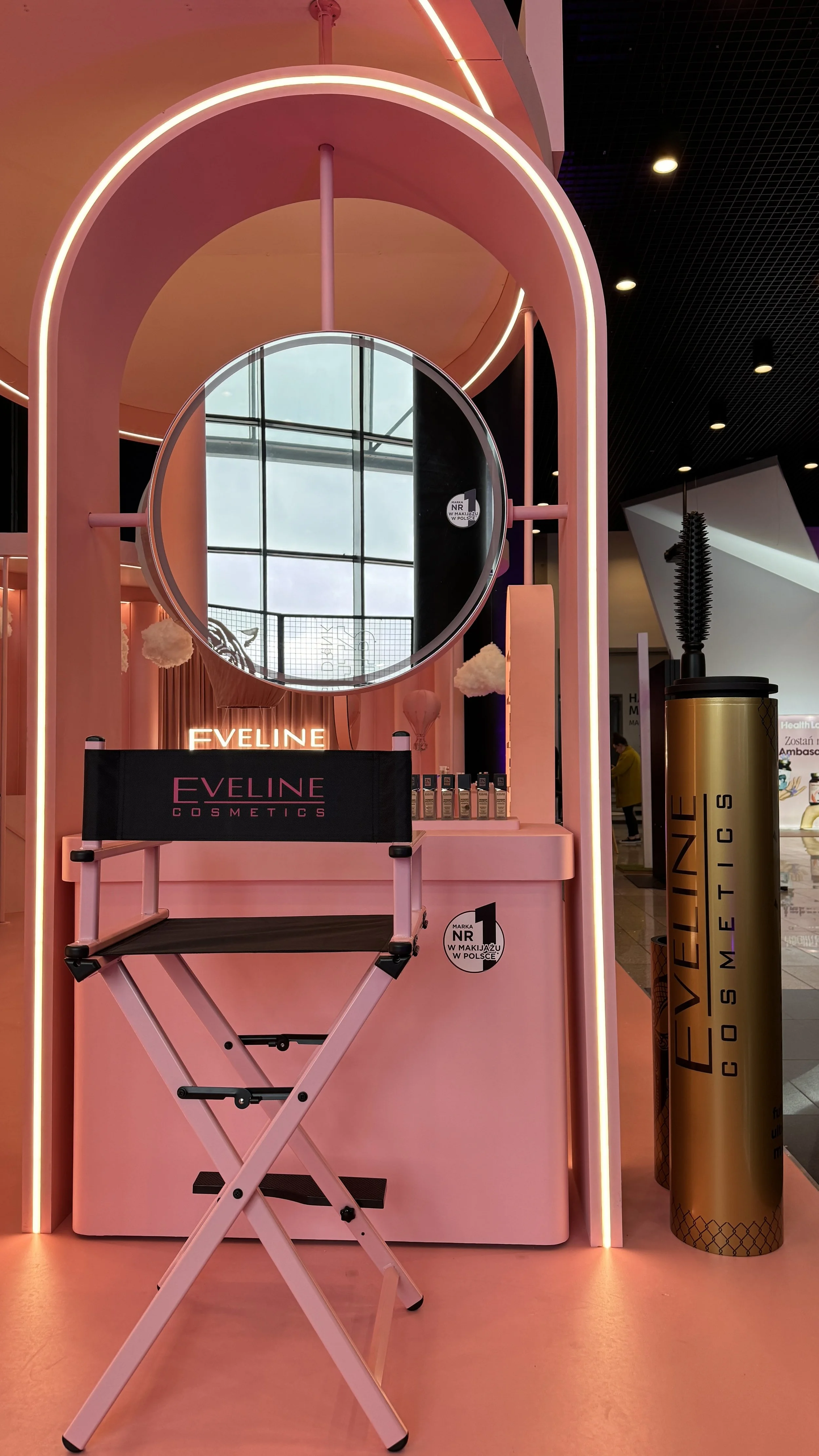 Pink makeup display booth with a circular mirror, a monitor with the text 'Eveline Cosmetics', a director's chair, and a cylindrical container with the text 'Eveline Cosmetics' on it, inside a shopping mall.