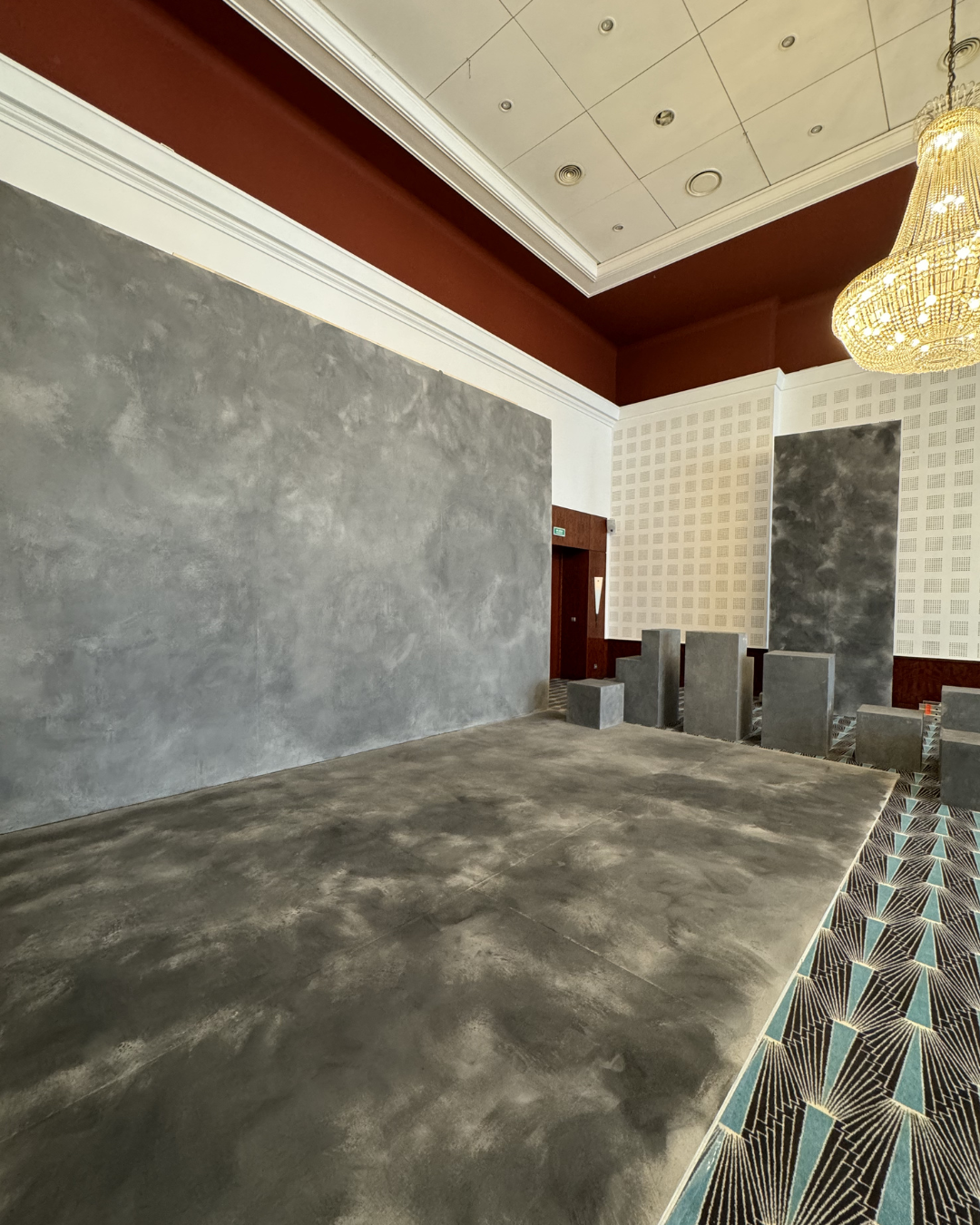 Empty hotel banquet room with polished concrete floors, a large textured gray wall, a white wall with a subtle geometric pattern, a crystal chandelier, and a carpet with an intricate geometric design.