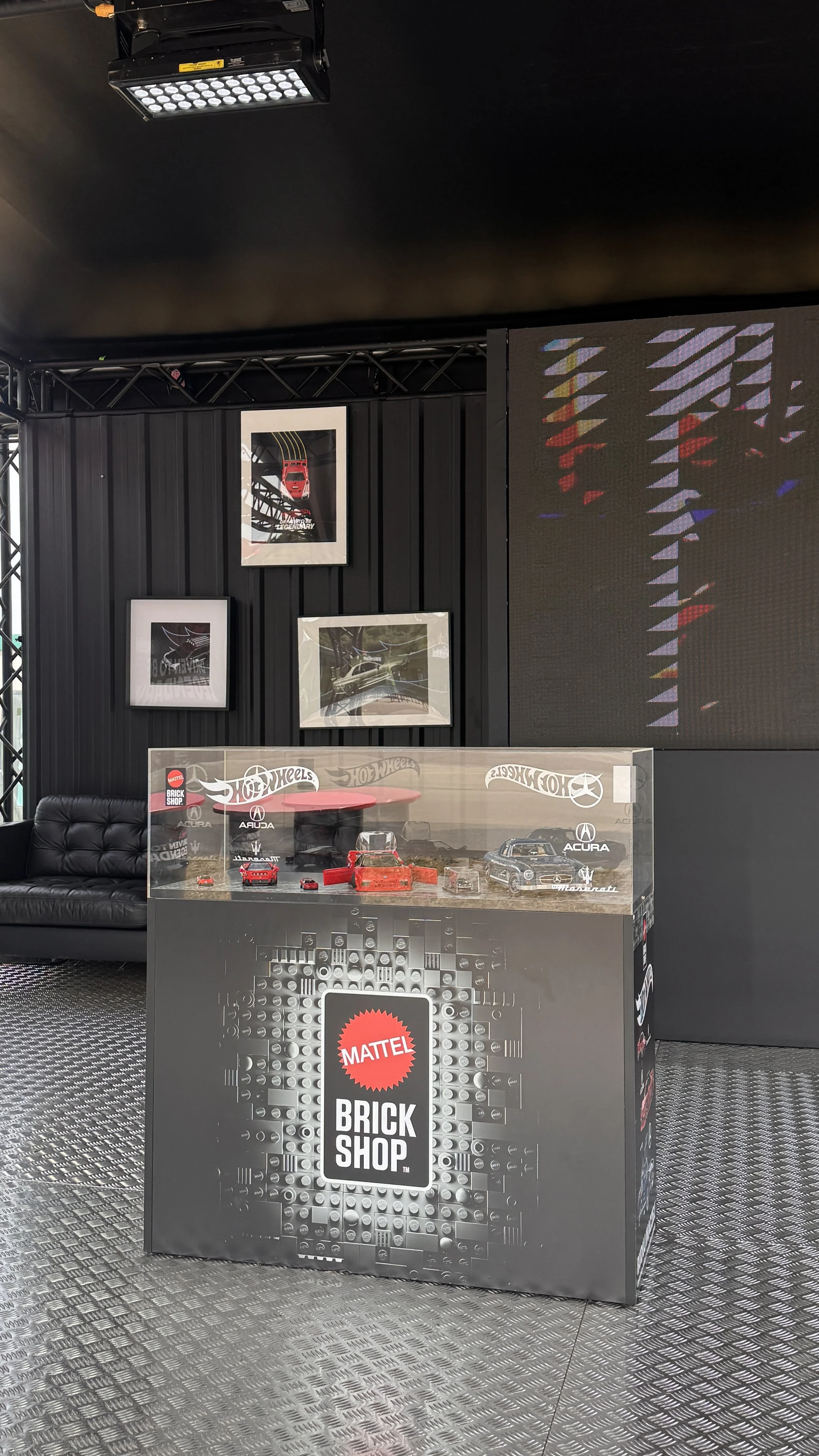 Display case in a Mattel Hot Wheels Brick Shop featuring toy cars, with black framed pictures of cars on a black wall behind and a black leather sofa to the side.