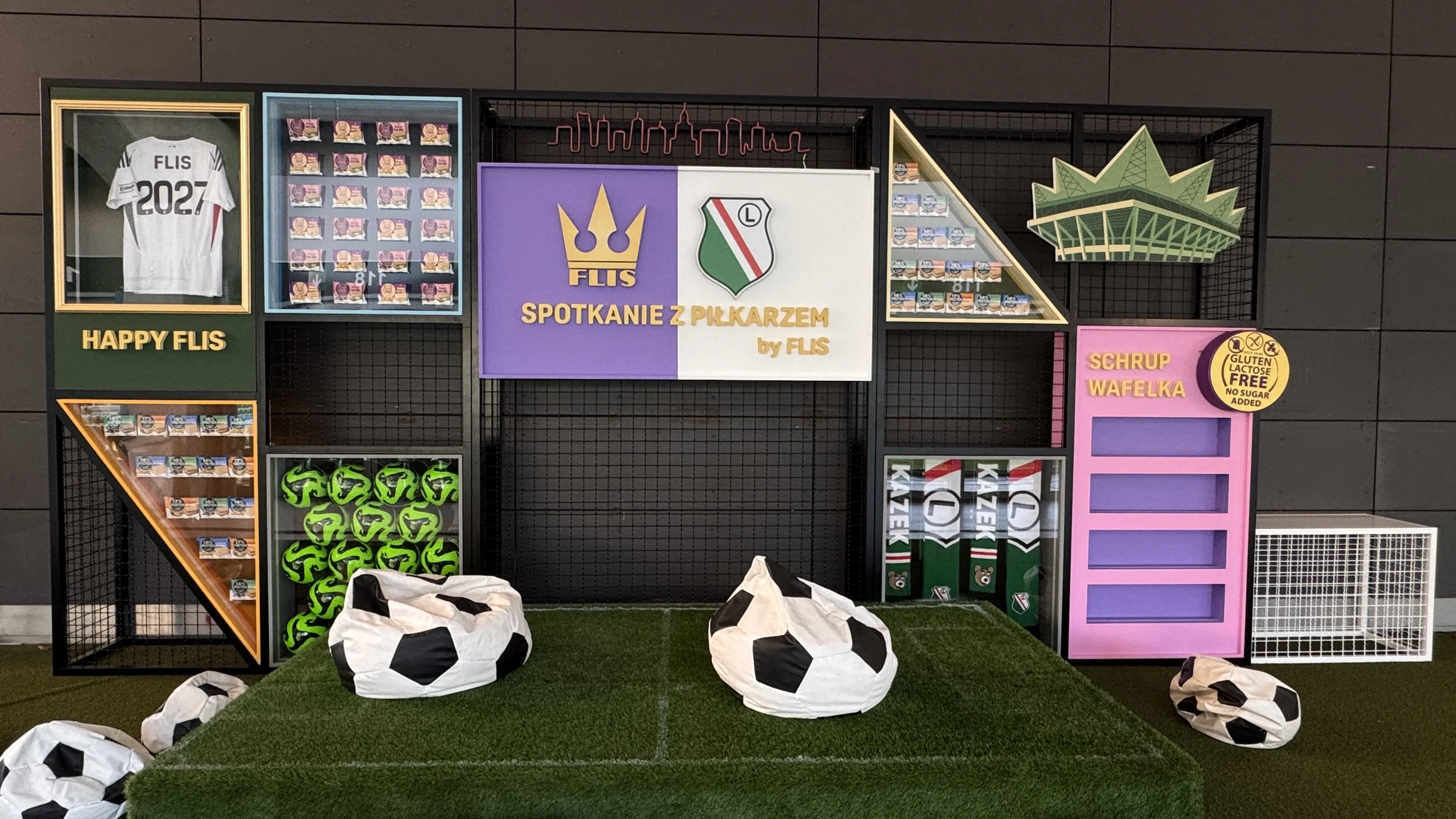 Decorative display with soccer-themed items including a white jersey with 'FLIS 2027' and a small logo, multiple stacks of snack packs, several green soccer balls, and beanbag chairs resembling soccer balls. There are also signs in Polish related to 