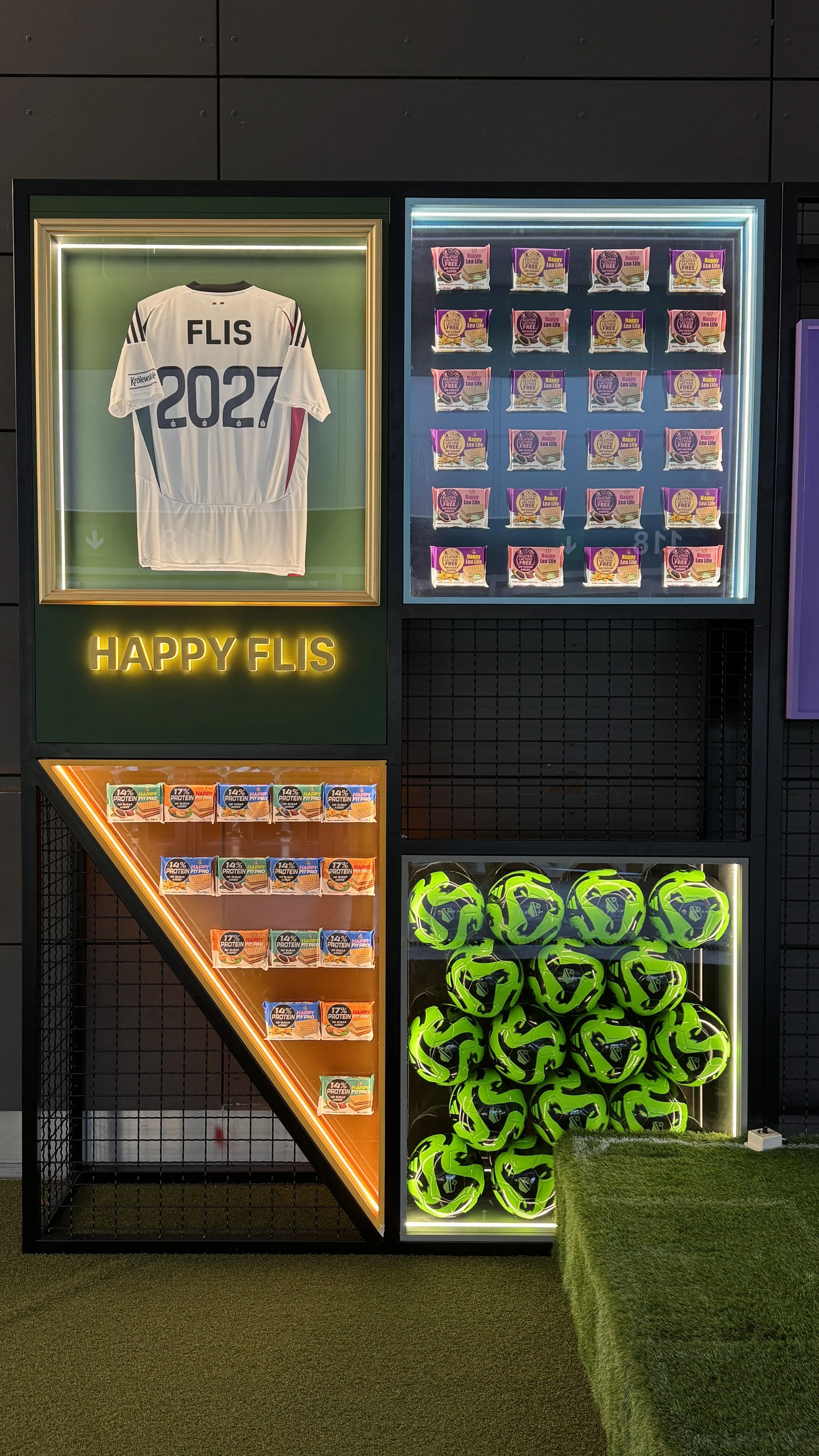 Display of a sports jersey, snack packs, and multiple neon green and black helmets at a merchandise stand. The jersey has 'FLIS 2027' on the back and the number 2027 printed below. The display includes a sign that says 'HAPPY FLIS'.