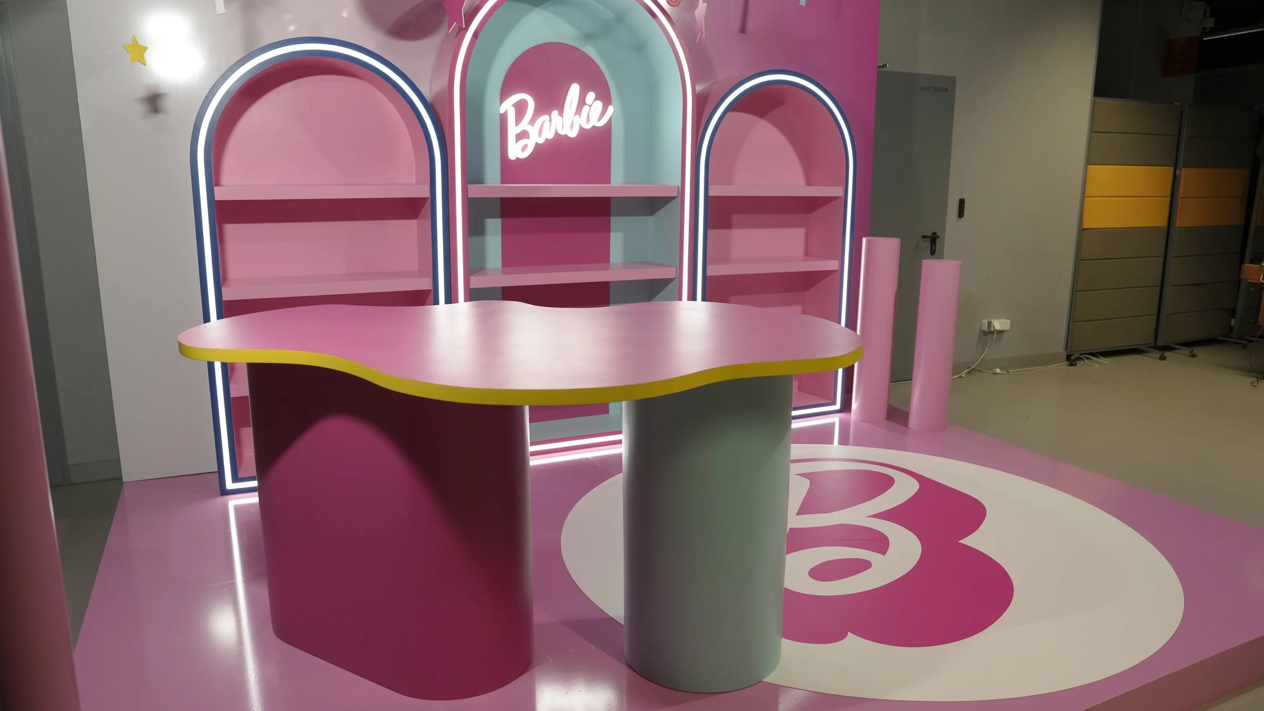 Pink and teal Barbie-themed display with shelves, a curved counter, illuminated arches, and Barbie logo, set on pink flooring with Barbie's logo.