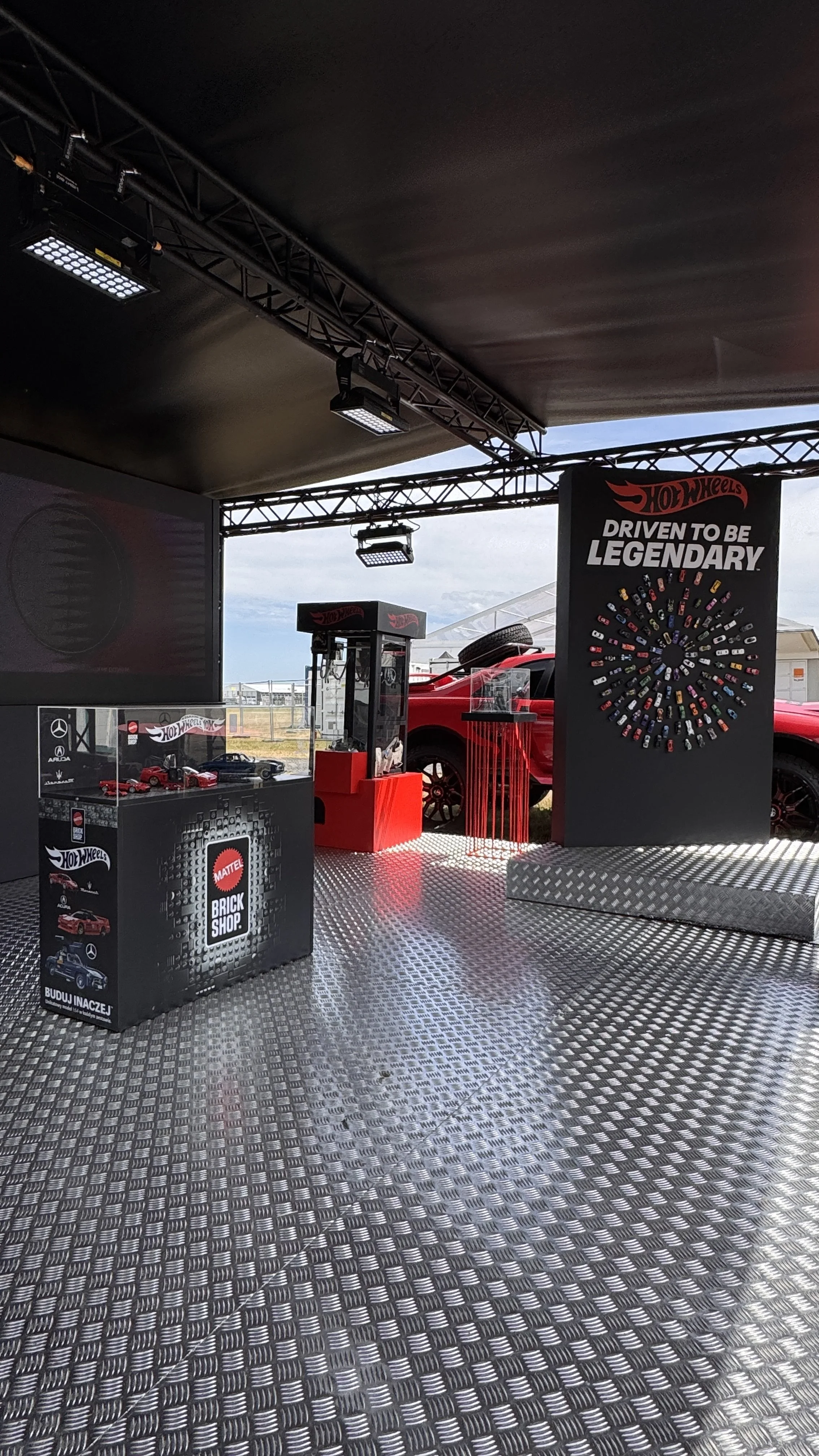 Hot Wheels display booth with a sign that says 'Driven to be Legendary,' a circle of miniature toy cars, a glass case with diecast model cars, and a smiling red toy car in the background. The booth has black and red elements with a textured metal flo