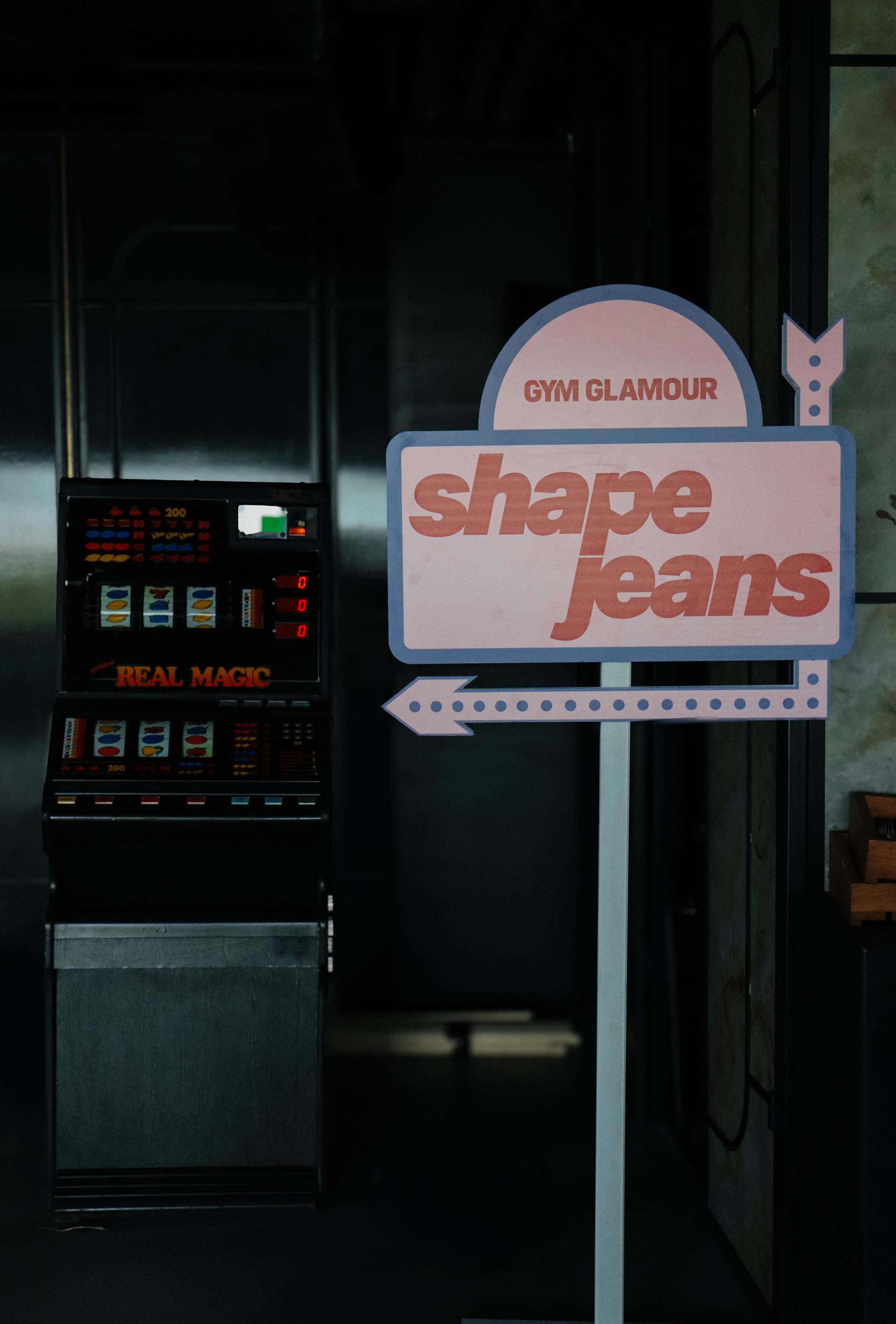 A sign with the text 'GYM GLAMOUR shape jeans' and an arrow pointing left, positioned outdoors near a slot machine, with a dark background.