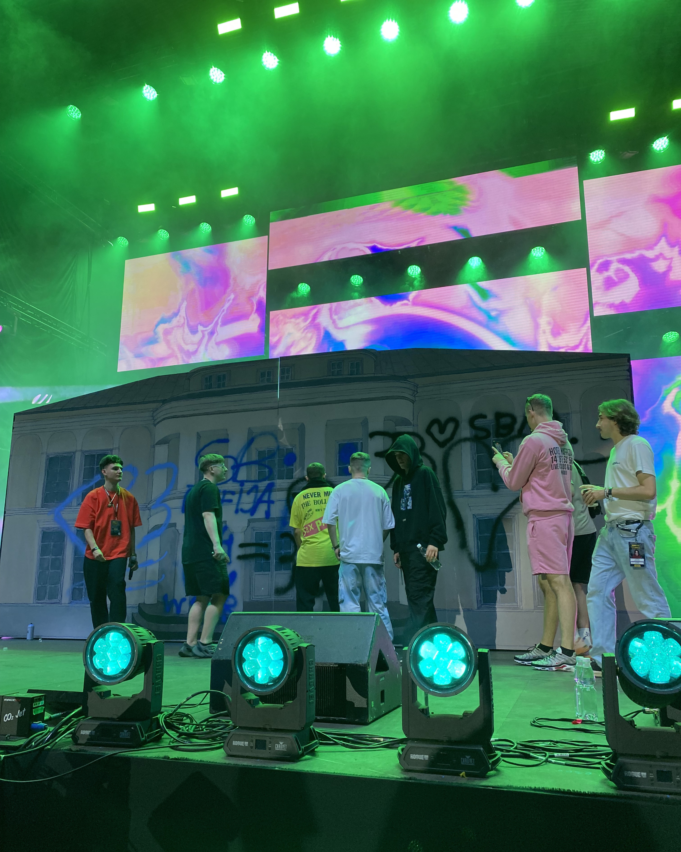 A group of people on a stage with colorful digital screens and green stage lights overhead. Some individuals are wearing casual clothes, and one person is holding a smartphone. There are stage lights in the foreground, and graffiti on the building be