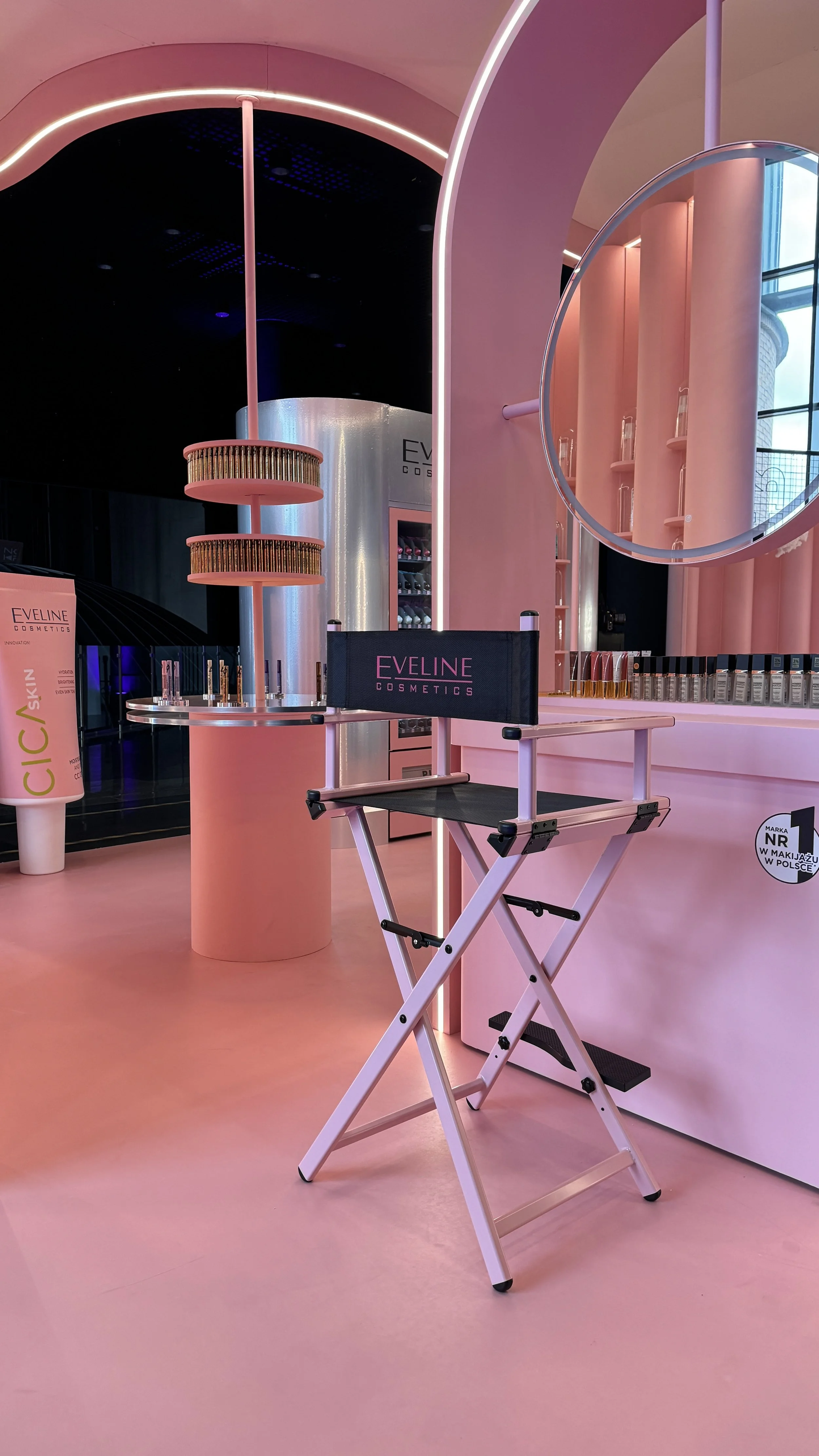 A makeup event setup with a pink theme, featuring a director's chair with 'Eveline Cosmetics' branding, a pink cylindrical display table with makeup brushes, and shelves with cosmetic products. The background includes pink curved walls and large wind