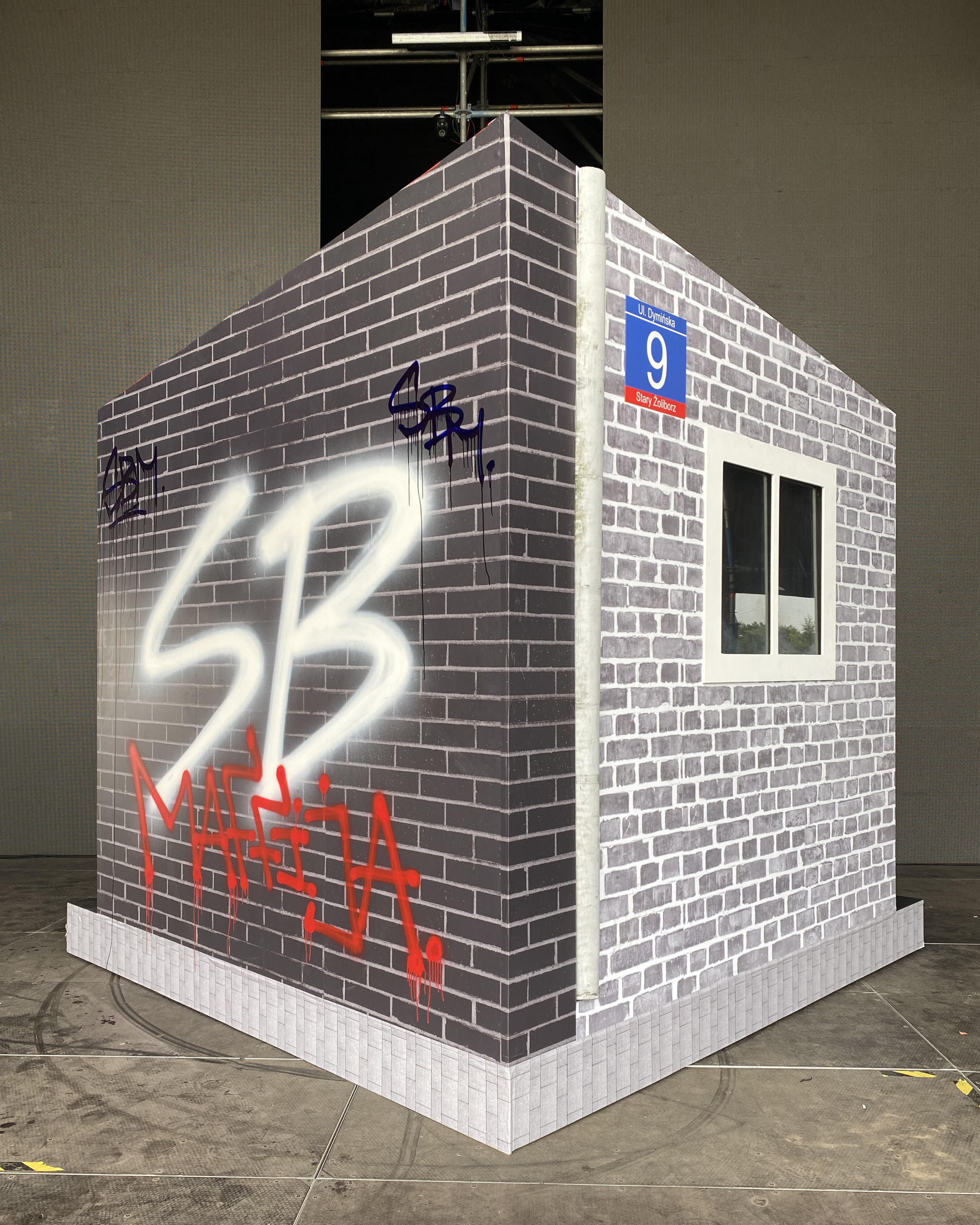 A large rectangular building or display with wallpaper looking like brick walls, with a window on the right side. There is graffiti in black spray paint on the bricks, and neon signs in white and red, with the text partially visible. The building is 